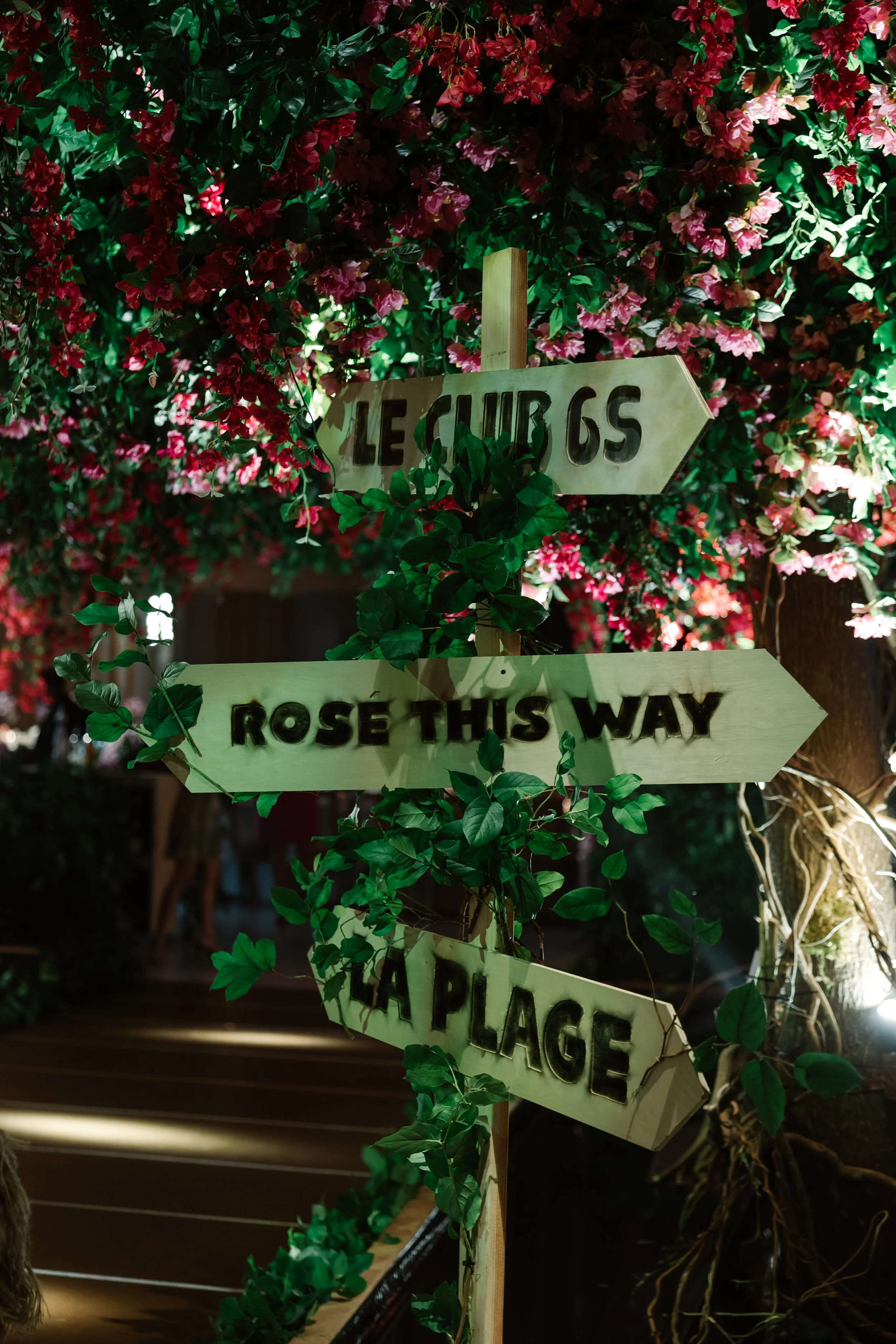 Wooden directional signs attached to a post amidst lush green leaves and pink flowers. The signs read 'LEISURE 65,' 'ROSE THIS WAY,' and 'PLACE.'