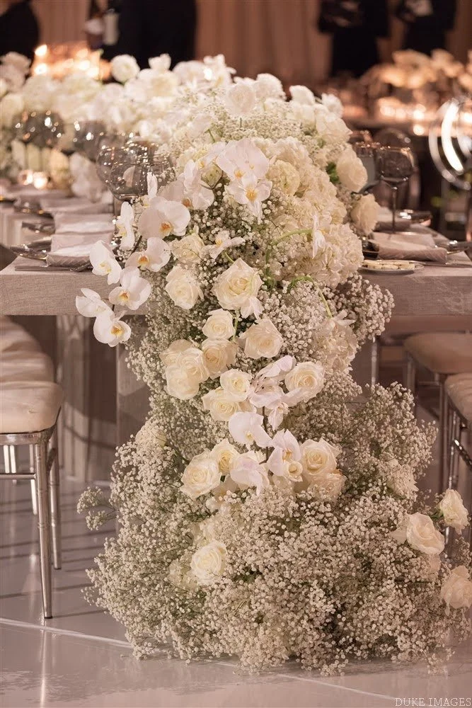 An elaborate floral centerpiece with white roses, orchids, and baby's breath arranged on a reception table at a formal event.