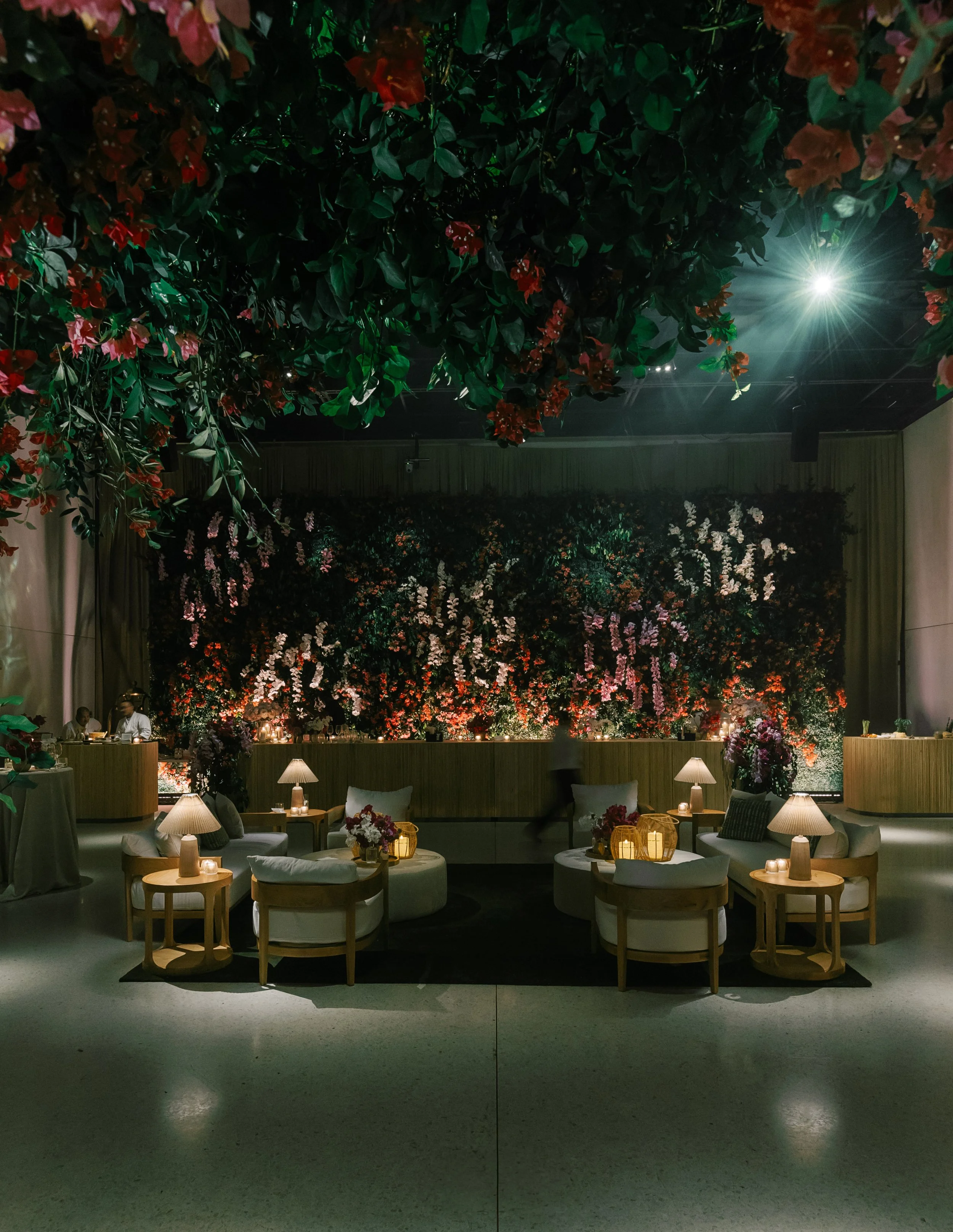 A dimly lit indoor venue with floral decorations on the wall and ceiling, featuring pink, red, and white flowers. There are small tables with lamps and floral arrangements, arranged around a seating area with white cushioned chairs and a black rug. I