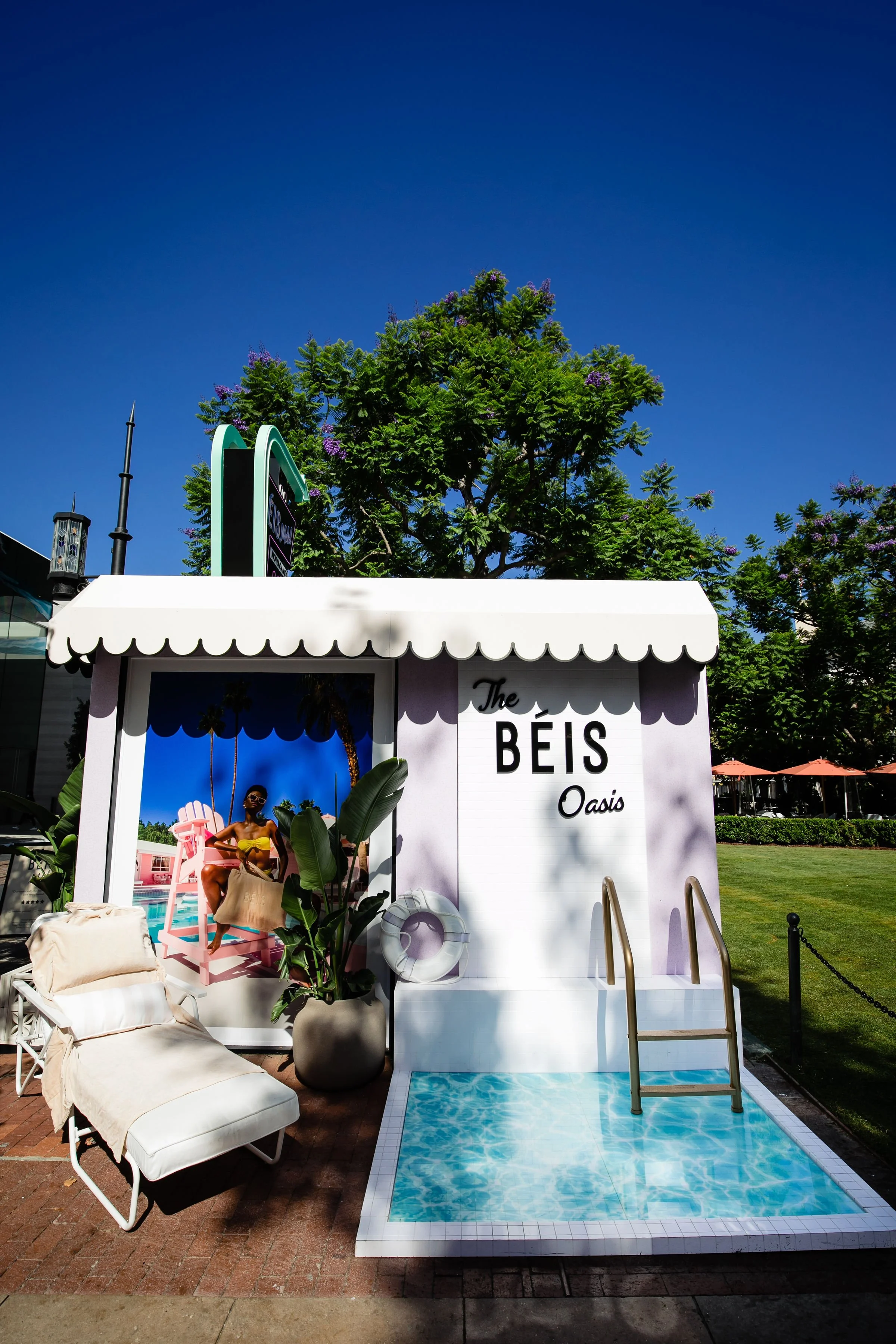 A marketing display for The BEIS Oasis featuring a small pool, lounge chair, tropical plants, and a large photograph of a woman in a yellow swimsuit sitting on a pink chair, with trees and a clear blue sky in the background.
