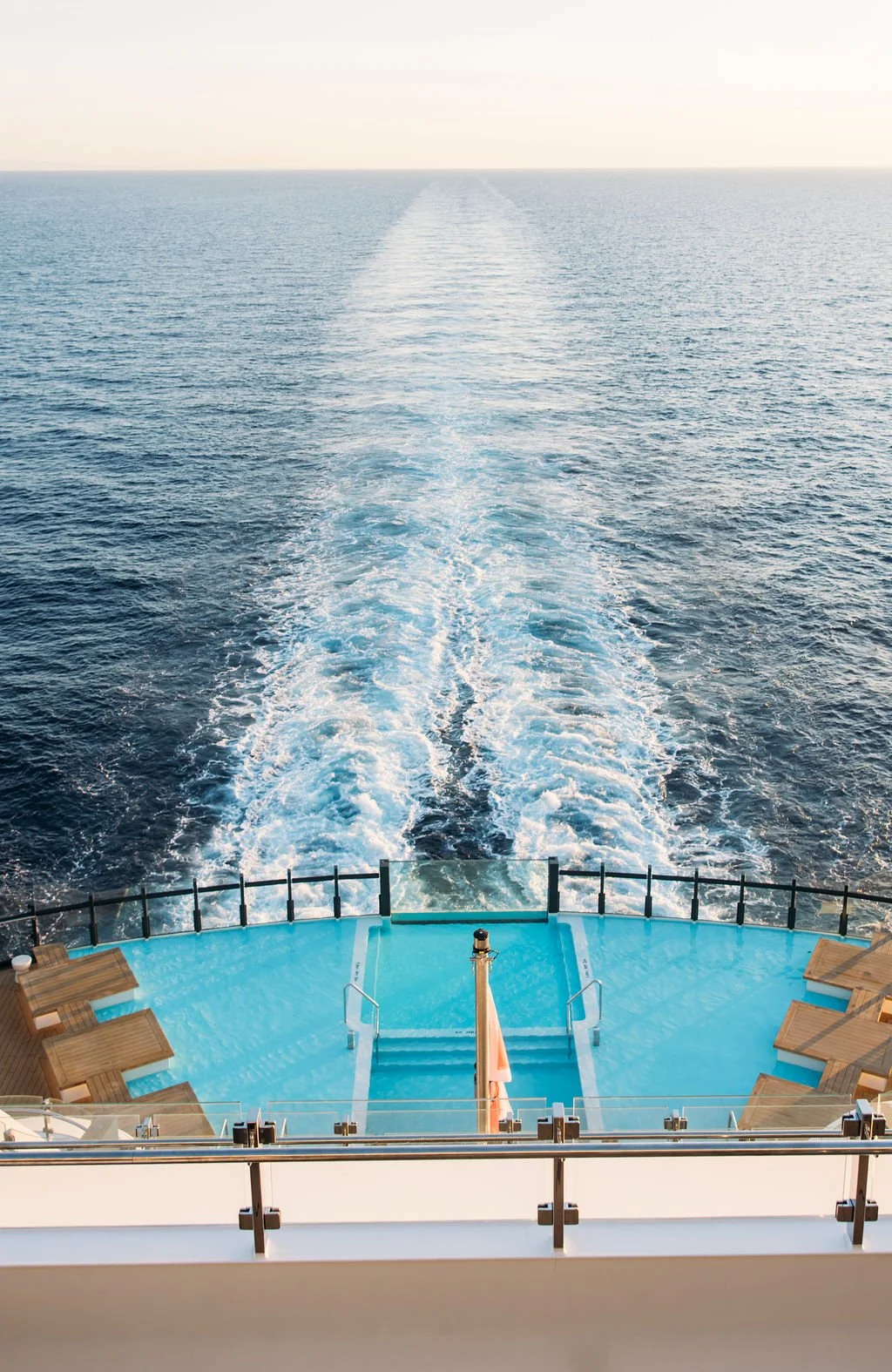 View from the back of a cruise ship showing the swimming pool and deck, with the ship's wake extending into the ocean on a clear day.
