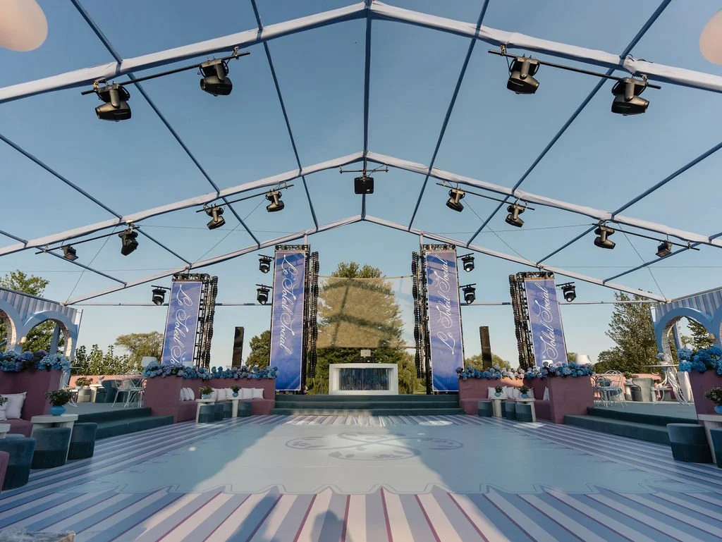 Empty outdoor event stage decorated with blue and pink flowers, large screens, and banners that read 'The Ball Room', with overhead string lights and trees in the background.