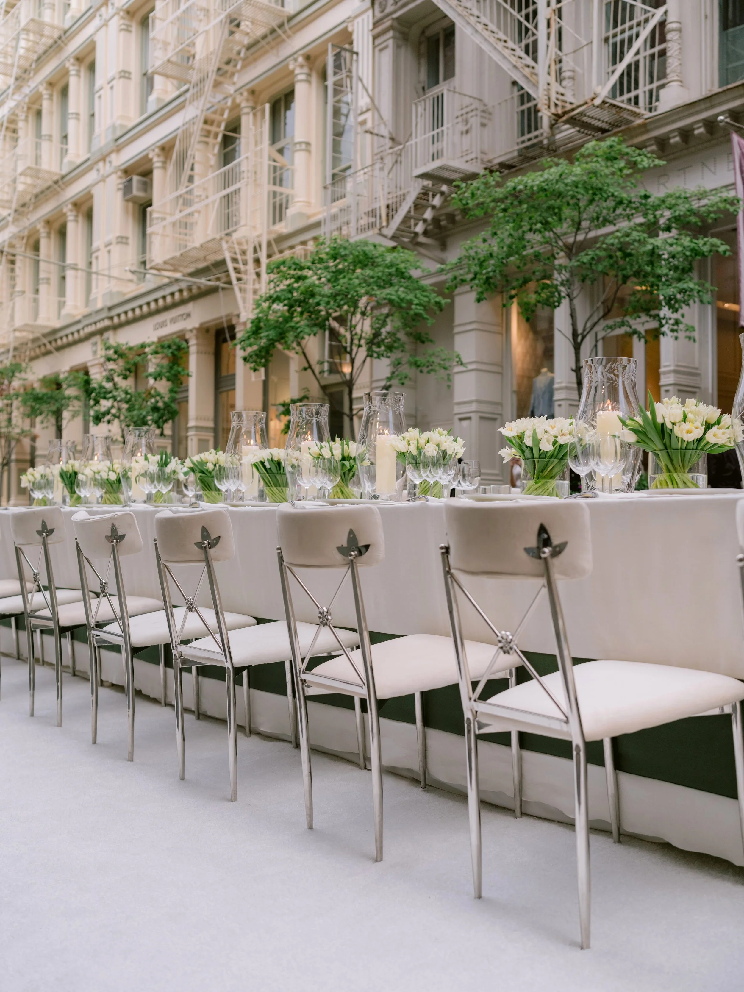 Elegant outdoor banquet table set with white chairs, floral centerpieces with white tulips, glass vases, and candles, in an urban street setting with historic buildings and trees in the background.
