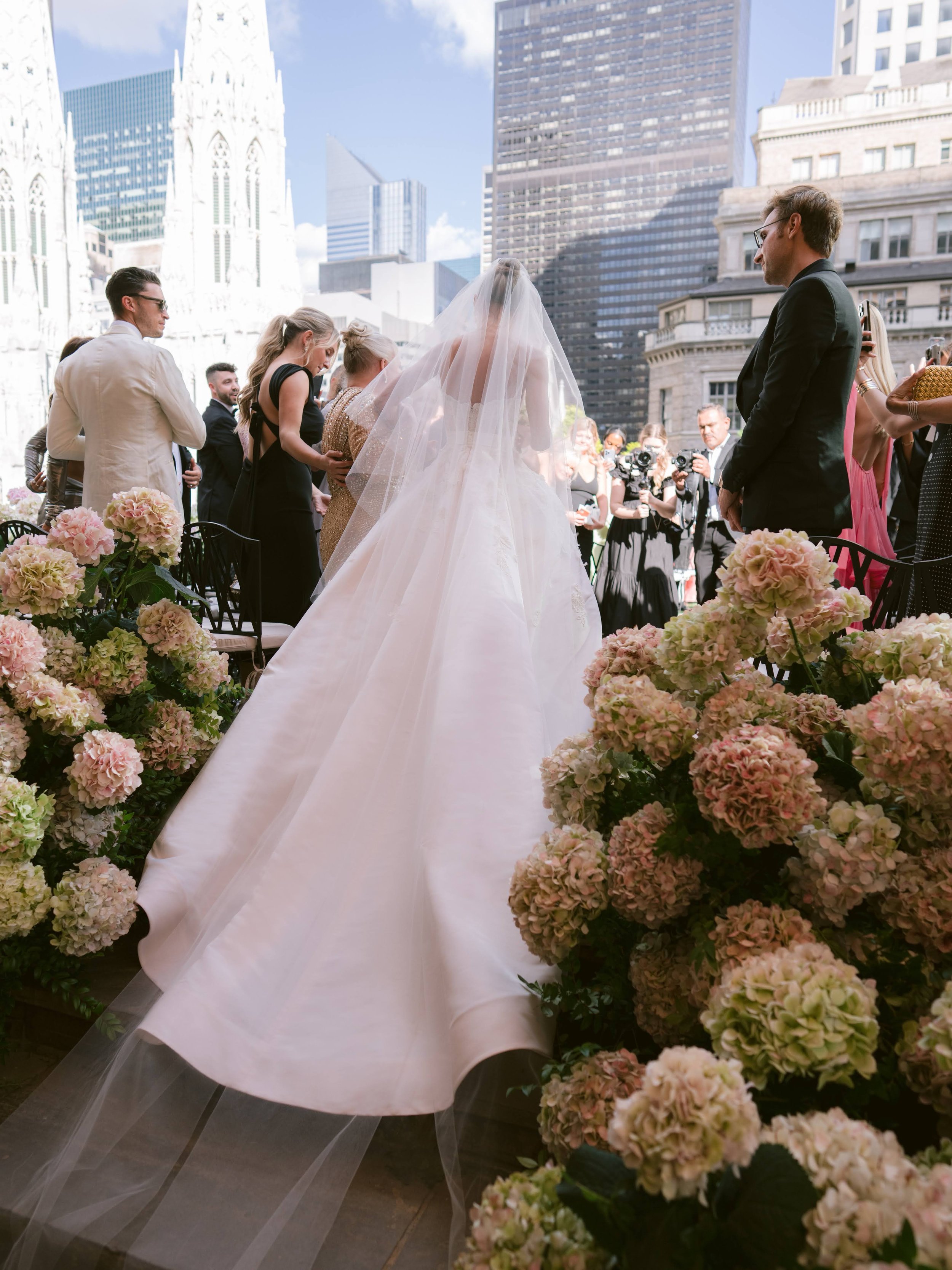 A bride in a white wedding gown and veil walking down the aisle at an outdoor wedding, surrounded by pink flowers and guests, with tall city buildings in the background.