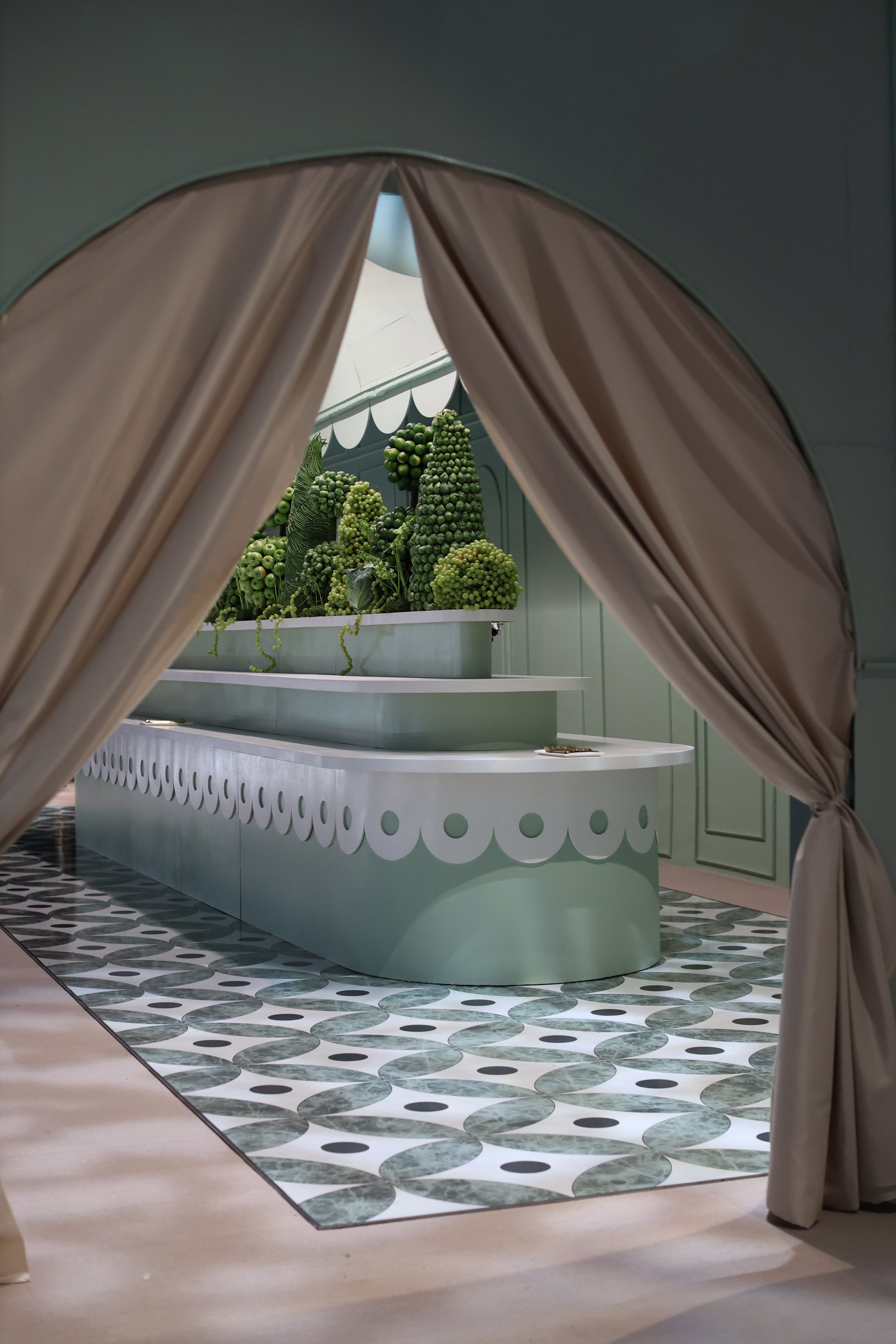 View through curtains of a display with green artichokes and vegetables on a decorative table in a pastel-colored space.