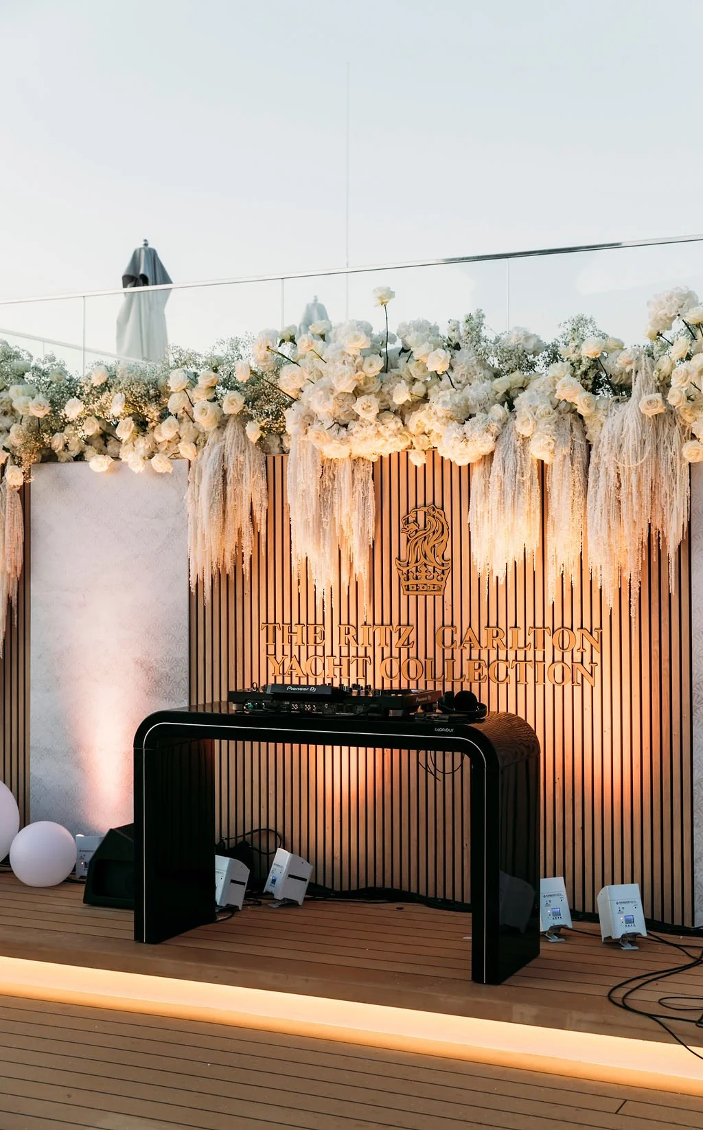 DJ setup on a wooden stage with a decorated backdrop featuring white flowers and the logo of The Ritz Carlton Yacht Collection.
