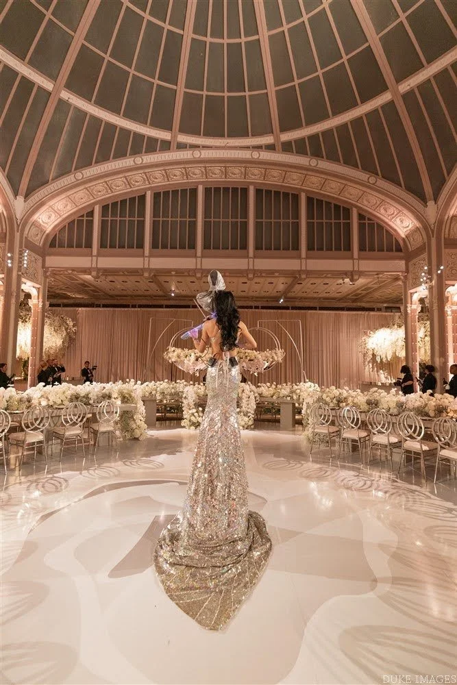 A woman in an elegant, sparkling gown stands on a polished dance floor in a grand, ornate ballroom with a high, domed ceiling. The room is decorated with white flowers and soft lighting.