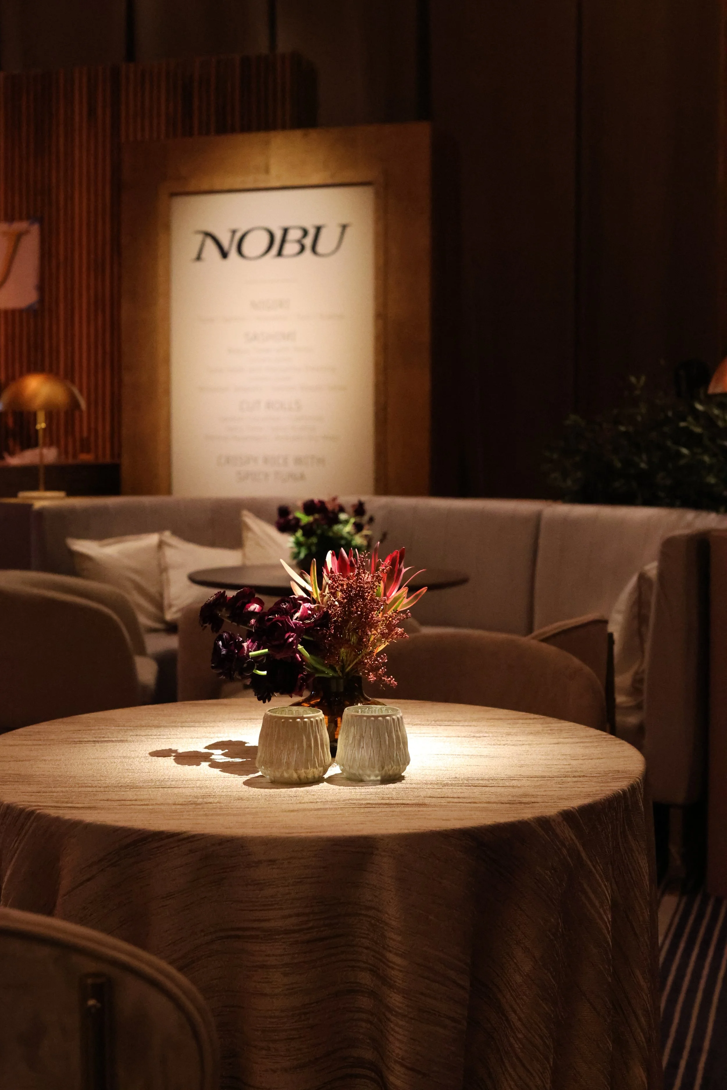 Elegant restaurant interior with a round table covered in a beige tablecloth, adorned with a floral centerpiece, and two small ceramic cups. In the background, there is a cushioned seating area and a framed menu on the wall.