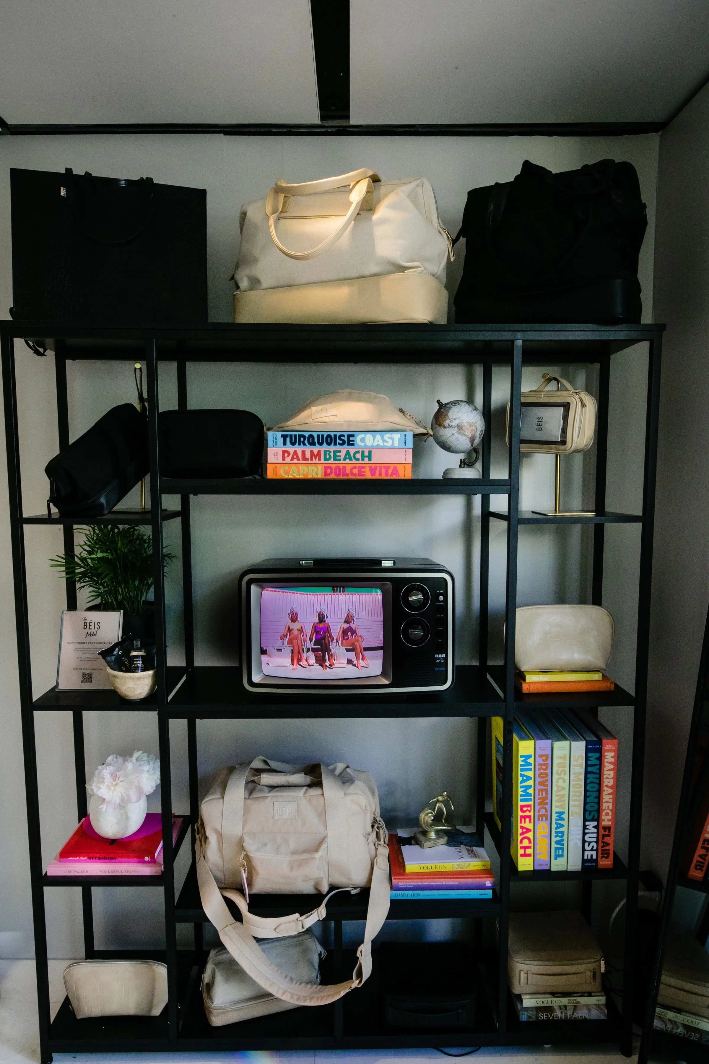 A black metal shelving unit with various bags on top, colorful travel books, a small globe, a vintage TV showing a dance scene, a potted plant, decorative items, and a collection of books. Some items include beige and black bags, a white decorative f