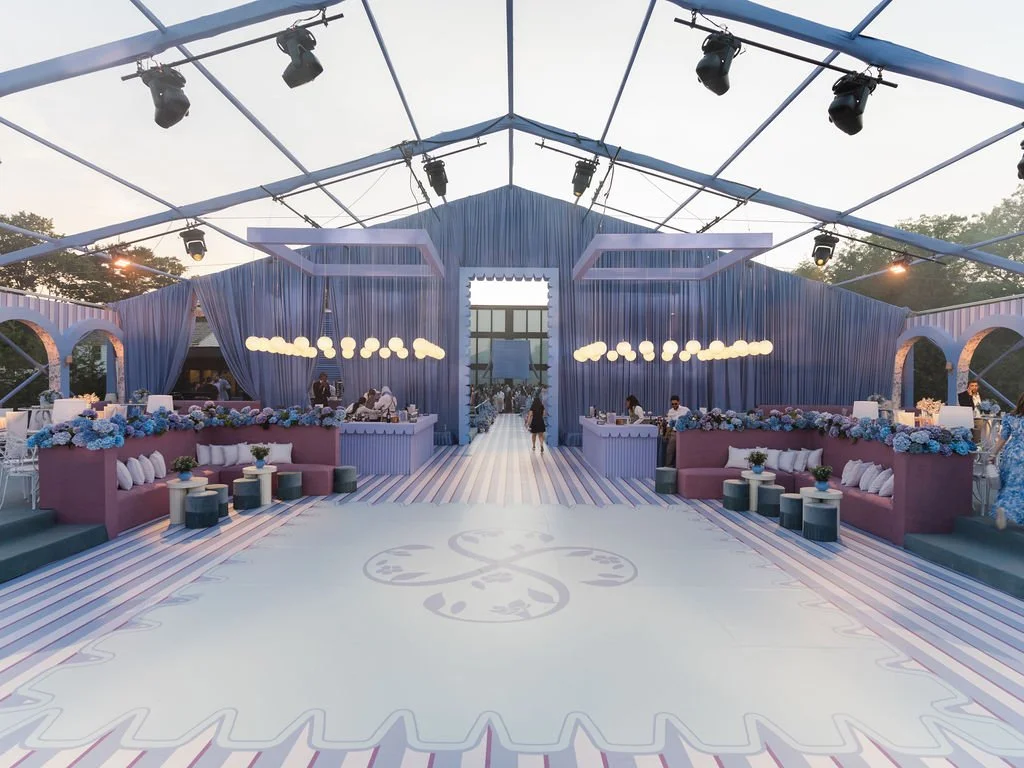 A decorated event space with blue drapes, pink couches, white tables, and floral arrangements, equipped with lighting and seating for guests.