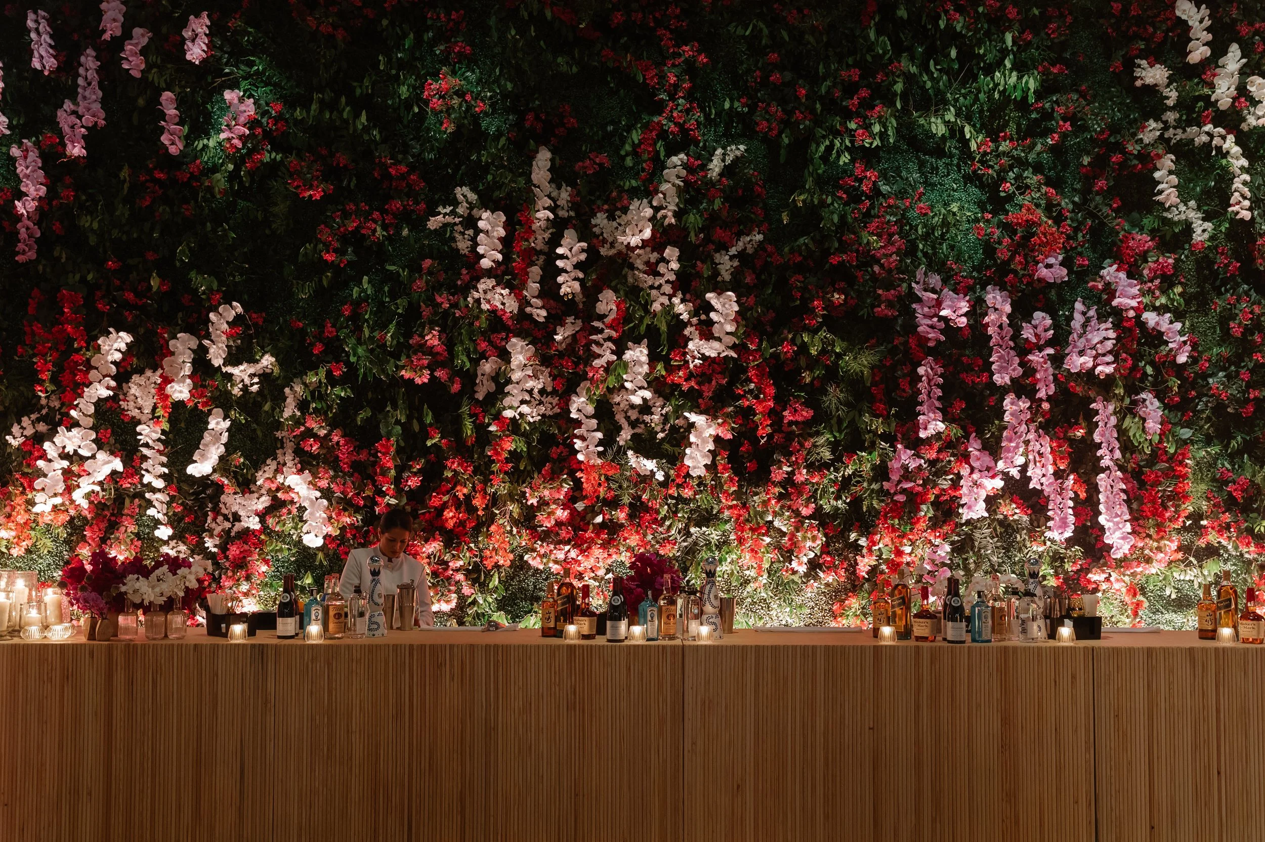 A bartender standing behind a bar with bottles and candles, set against a wall decorated with a large, vibrant floral arrangement of pink, red, and white flowers.