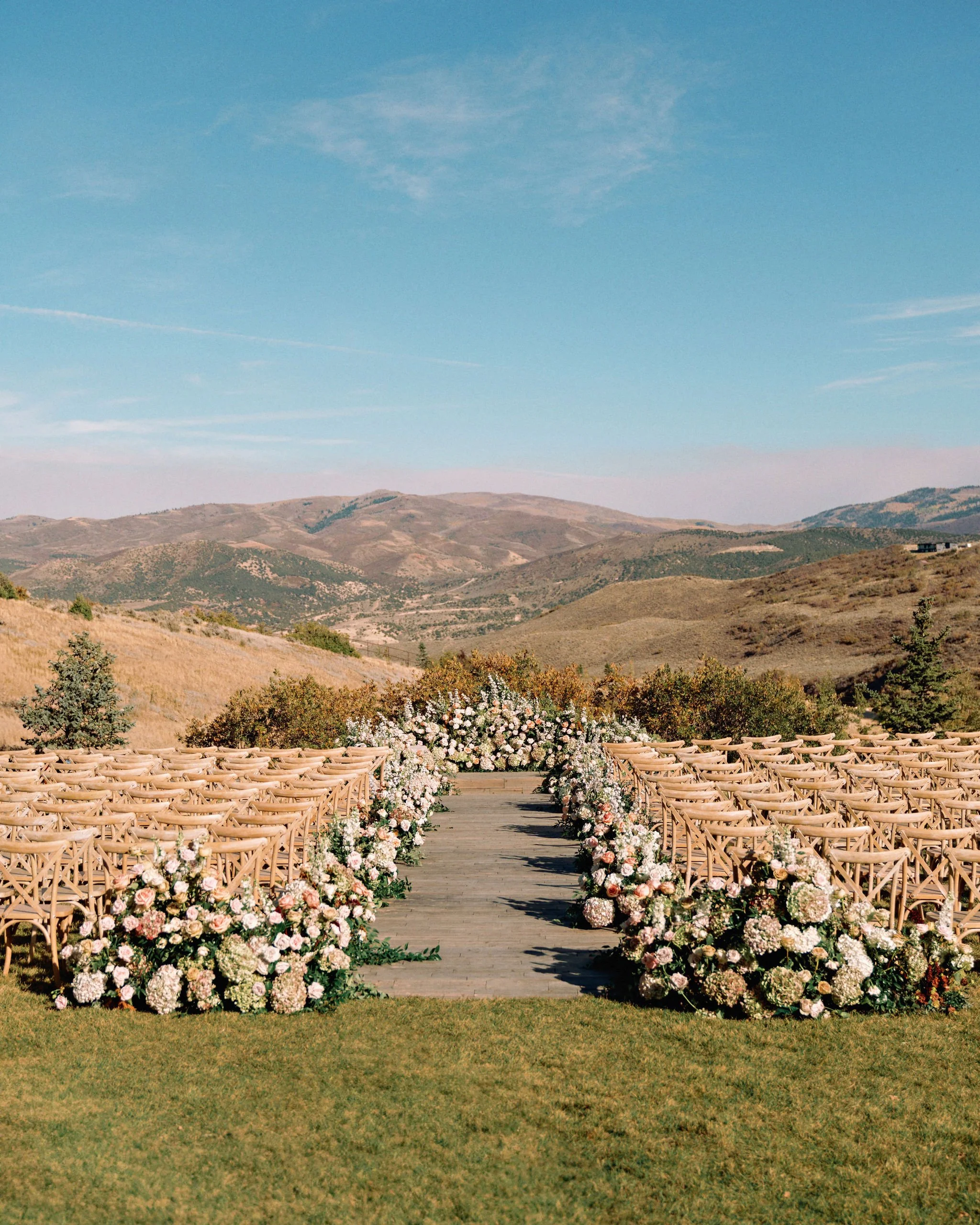 Outdoor wedding setup with rows of wooden chairs and floral arrangements along a wooden aisle, set against a scenic landscape of rolling hills and a blue sky.