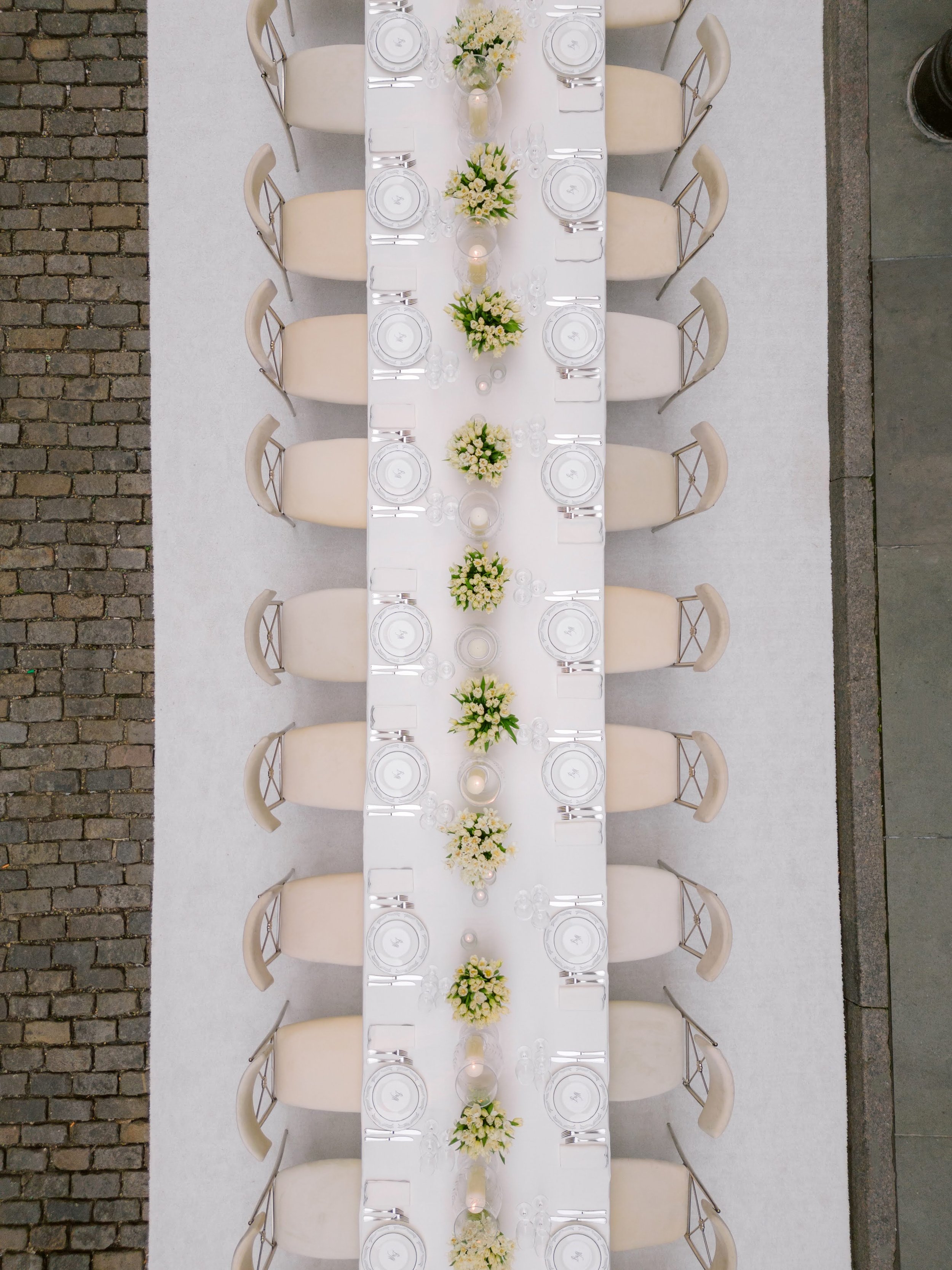 Overhead view of a long banquet table set for a formal event with white tablecloth, floral centerpieces, candles, and place settings, on a sidewalk with brick and concrete paving.