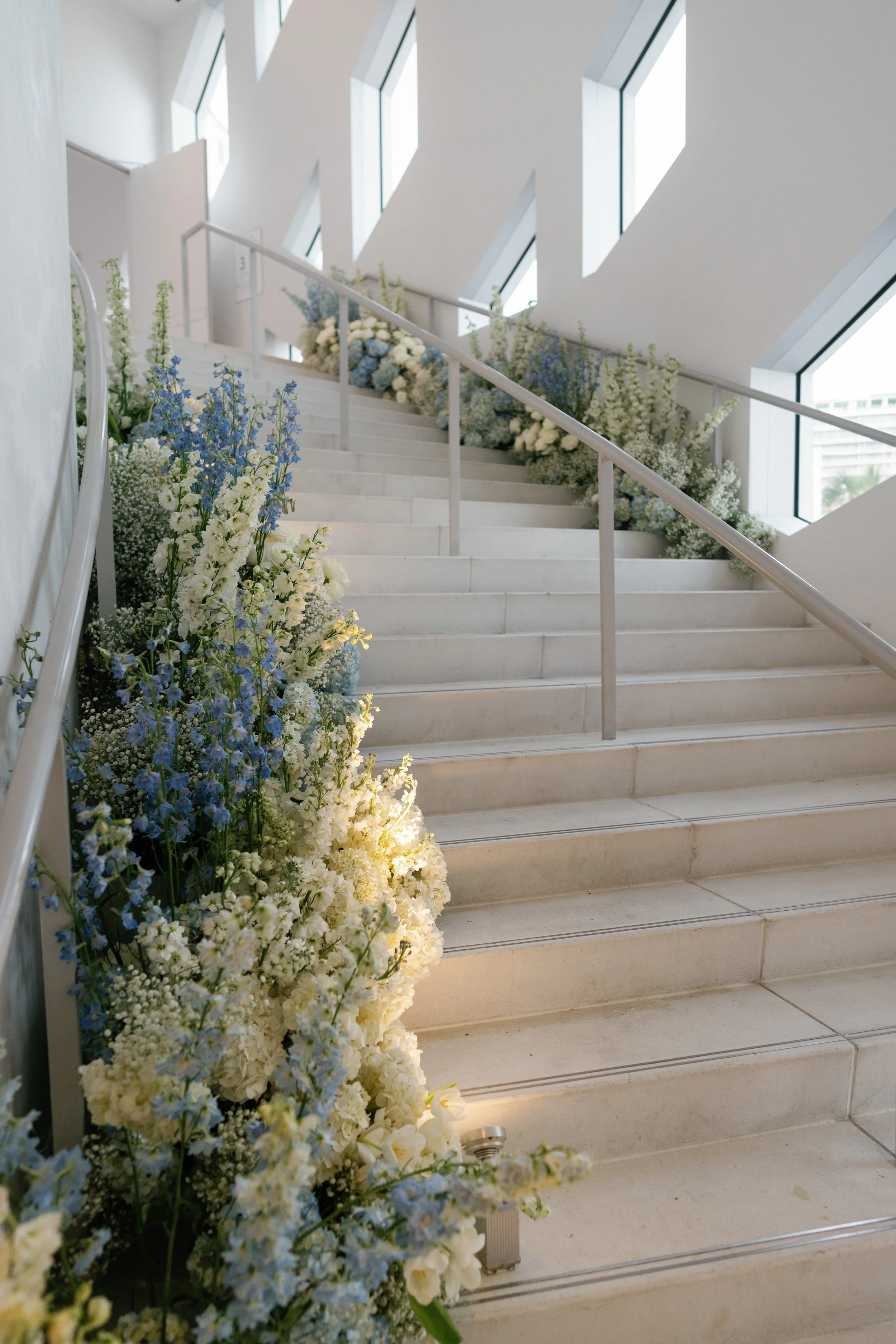 Indoor staircase decorated with white and blue flowers along the side railing, with large windows providing natural light.