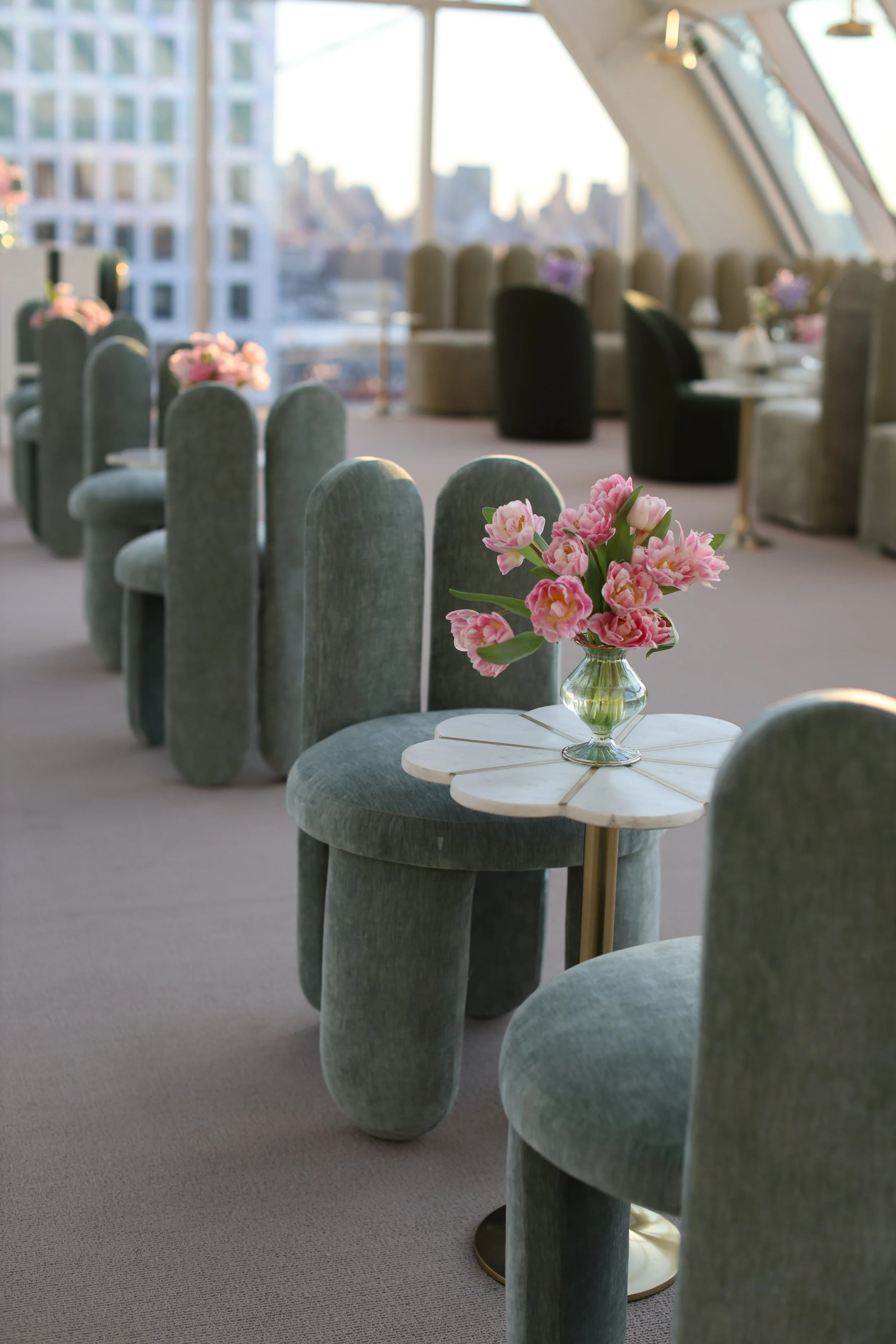 Indoor lounge area with pastel green velvet chairs, a marble-topped small table with a pink flower arrangement, large windows with city skyline view, and soft natural lighting.