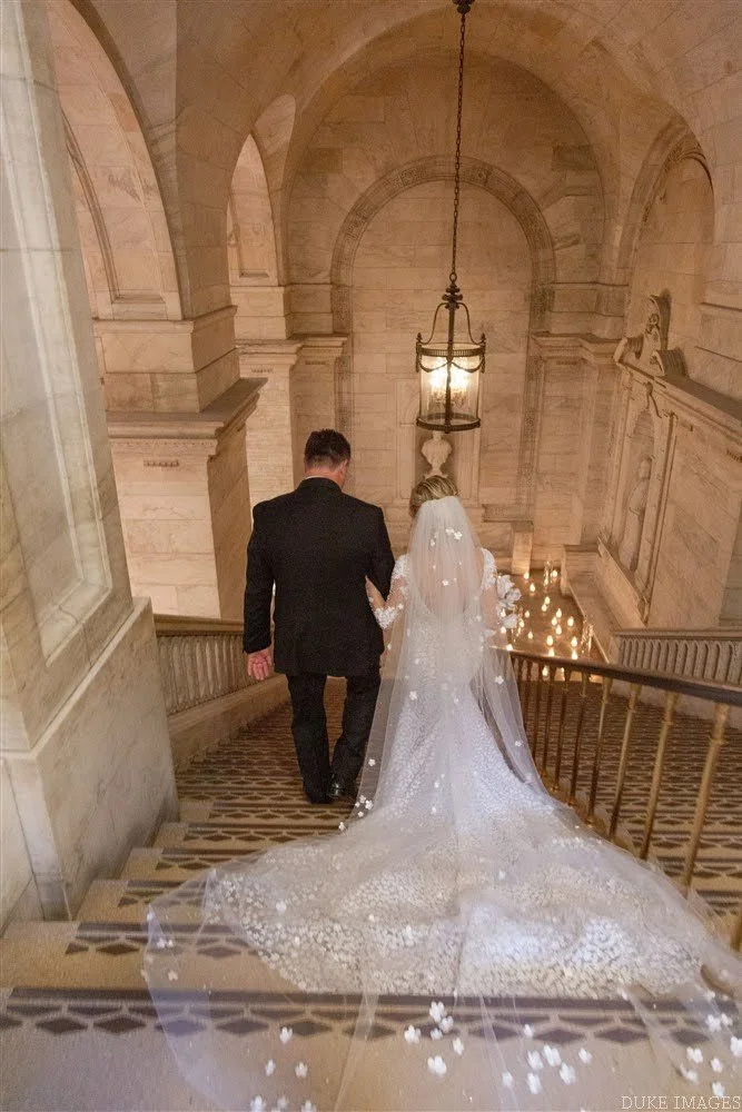 Bride and groom walking down a grand marble staircase in a historic building with arches and a hanging lantern, with the bride wearing a wedding dress with a long train and veil.