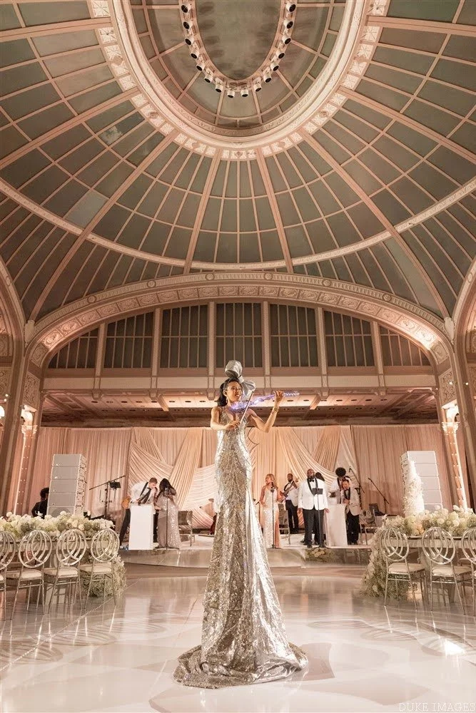 A woman dressed in a glamorous silver gown plays the violin on an elegant stage in a luxurious ballroom with high vaulted ceiling and ornate decor, with a live band performing in the background.