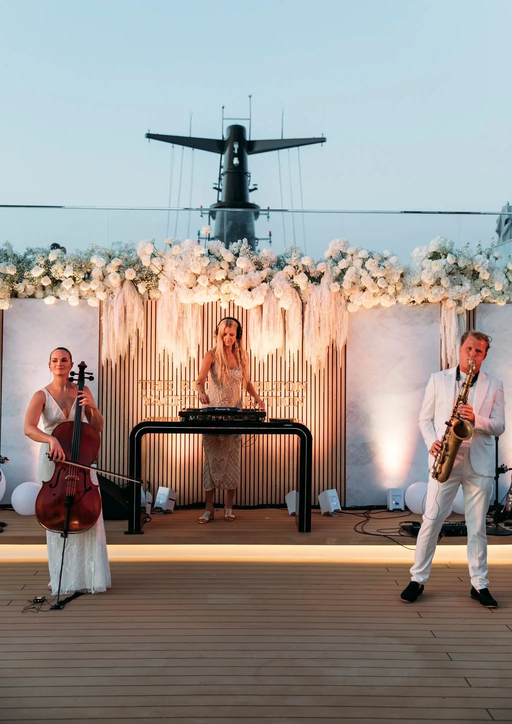 A live band performing on an outdoor stage decorated with white flowers and drapes, with a helicopter in the background at dusk.