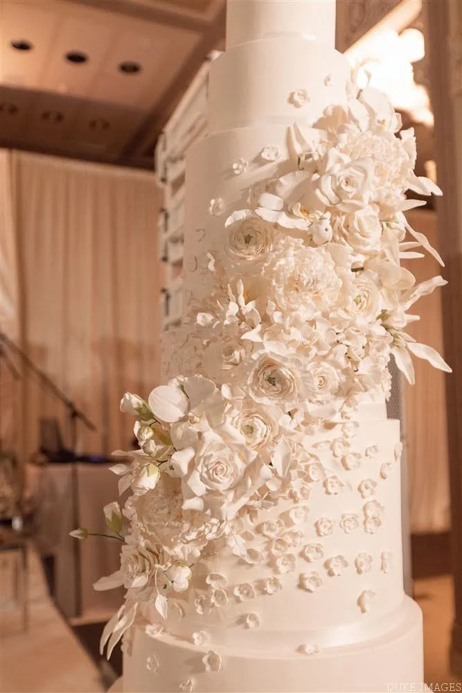 White wedding cake decorated with intricate white flowers and petals cascading down the side, set in a warmly lit indoor venue.