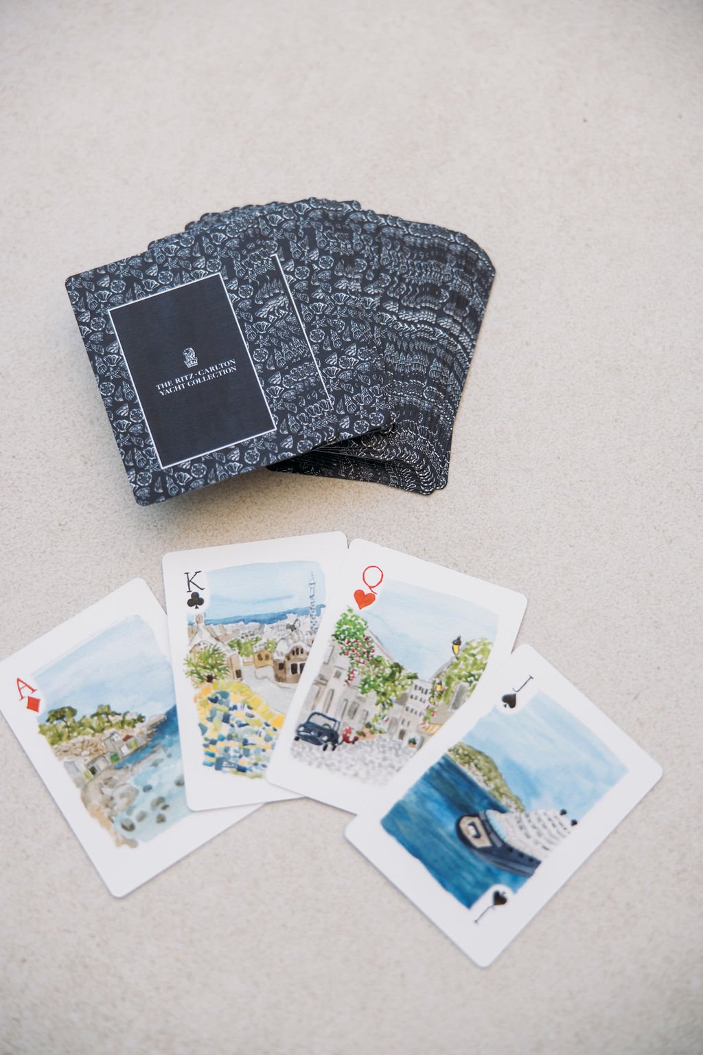 A deck of cards from The Ritz-Carlton Yacht Collection with five drawn cards showing scenic illustrations, including seaside views, a cityscape, and a harbor.
