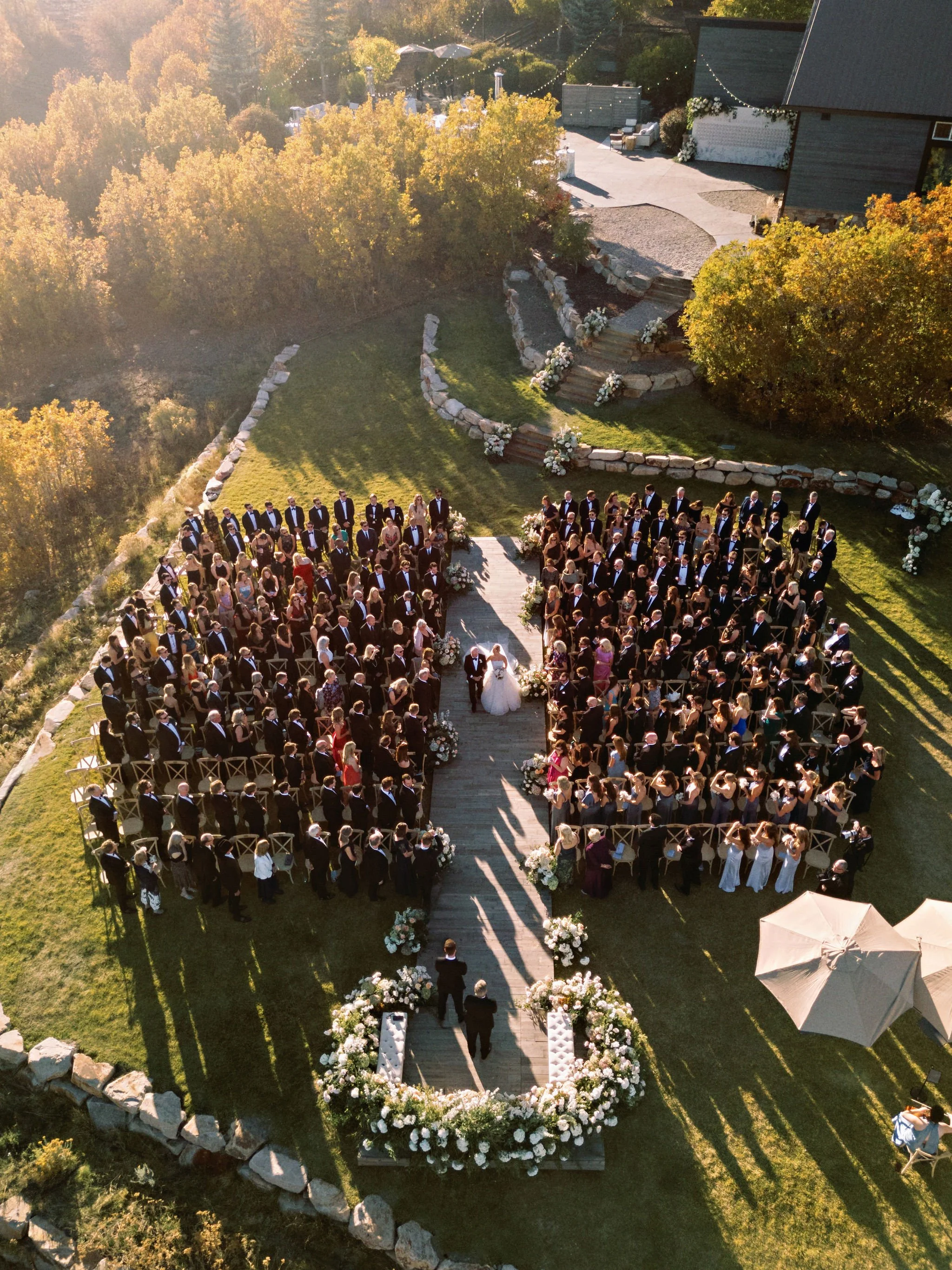 A wedding ceremony taking place outdoors in a grassy area, with a bride and groom standing under a floral arch at the altar, surrounded by seated guests. The setting is decorated with flowers and greenery, and there are trees and a building in the ba