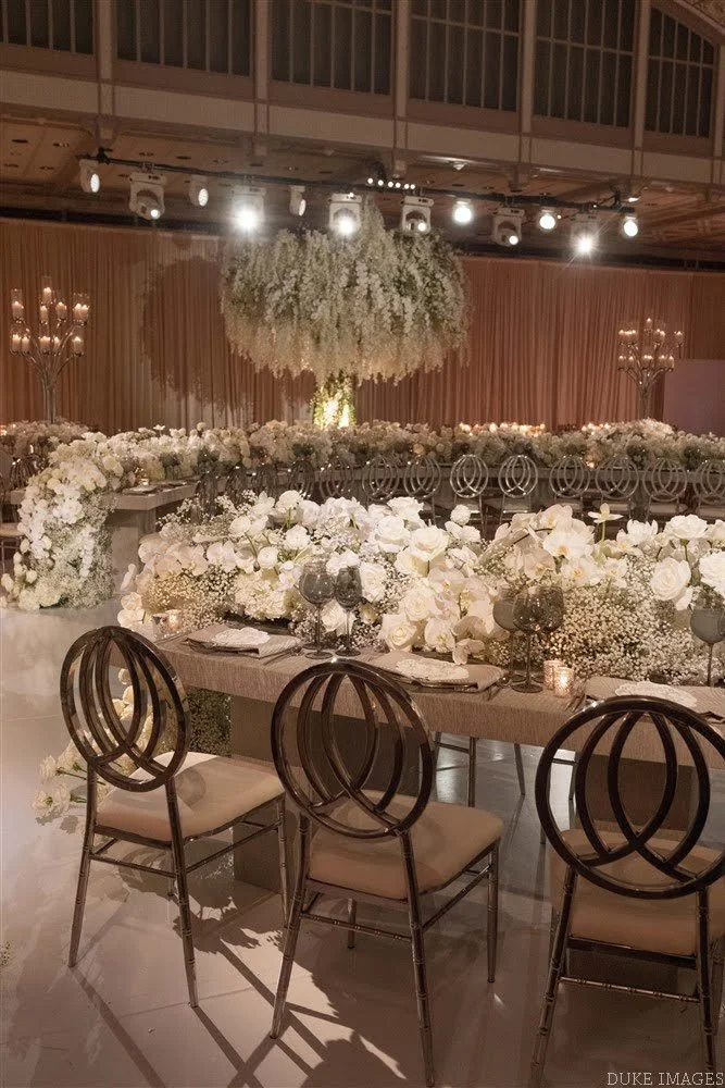 Elegant banquet hall decorated with white flowers and candles, with a large floral chandelier hanging from the ceiling and rows of tables with floral centerpieces and candlelit décor.