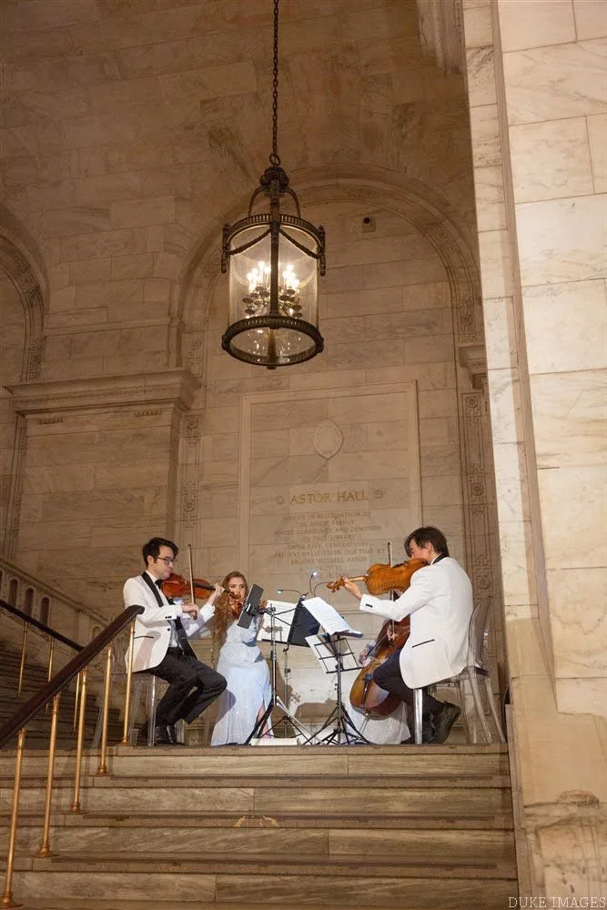 Three musicians performing in an elegant indoor space with marble walls and a large chandelier, two playing violins and one on a cello, with music stands in front of them.