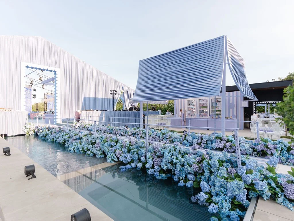 A modern outdoor venue decorated with blue and white hydrangeas, featuring a small blue-roofed building and a large white tent in the background, set up for an event or celebration.