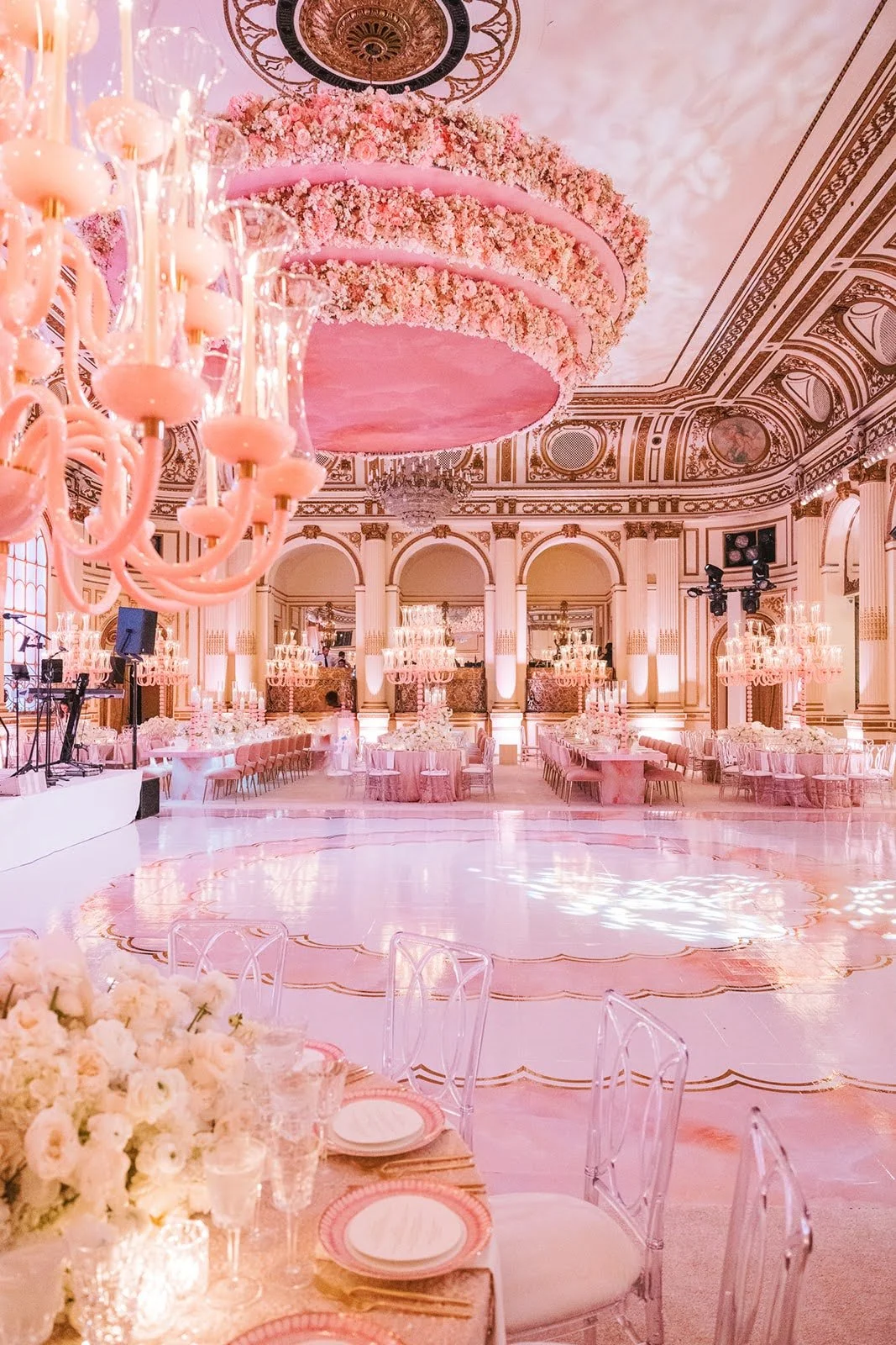 A grand ballroom decorated in pink and gold for a wedding or event, with floral chandeliers, ornate ceiling details, and elegant tables with pink linens and floral centerpieces.