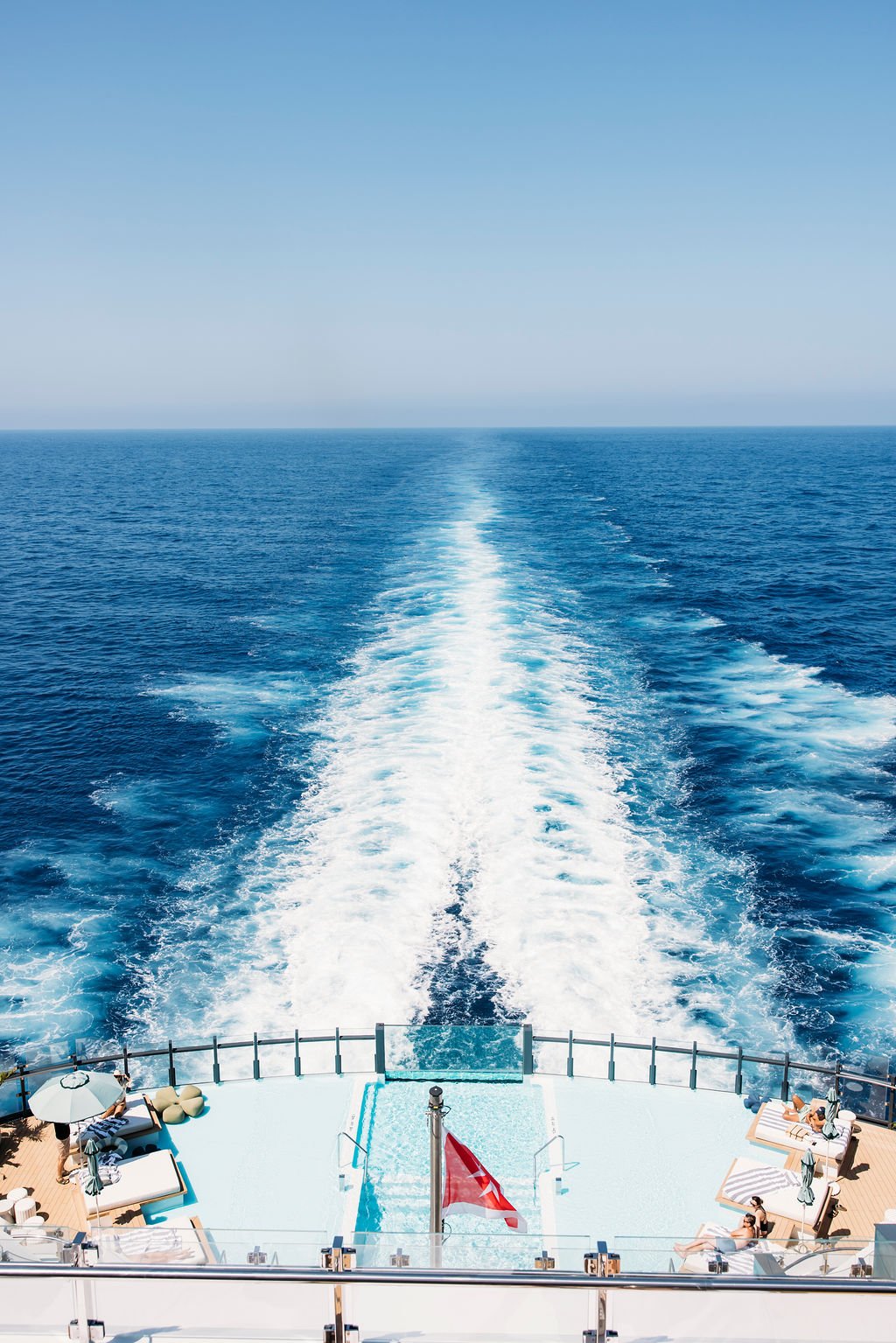 View from a cruise ship showing the ocean and the ship's stern with a swimming pool and lounge chairs.