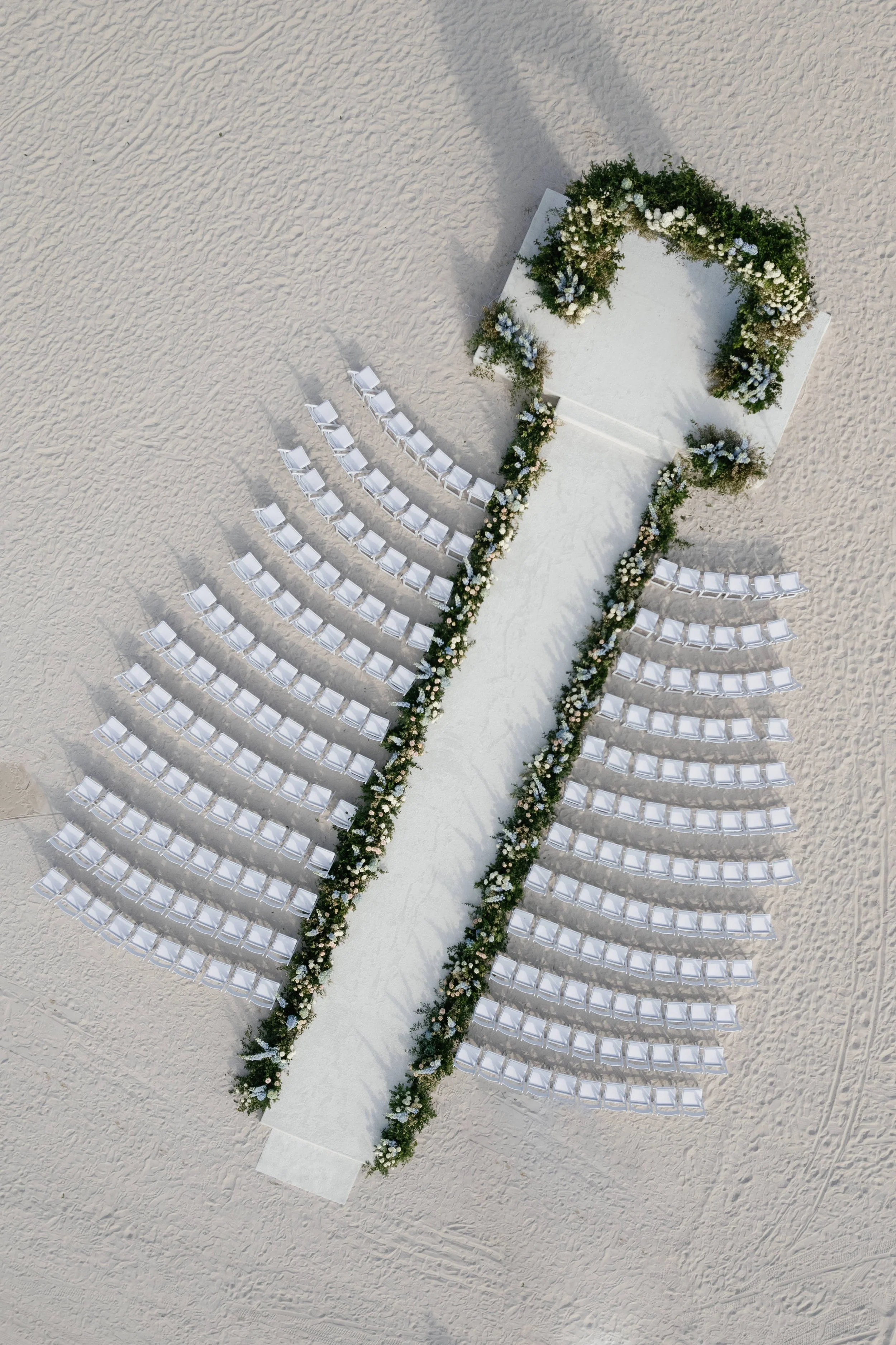 Aerial view of a wedding aisle on a sandy beach, decorated with flowers, with rows of white chairs on either side.