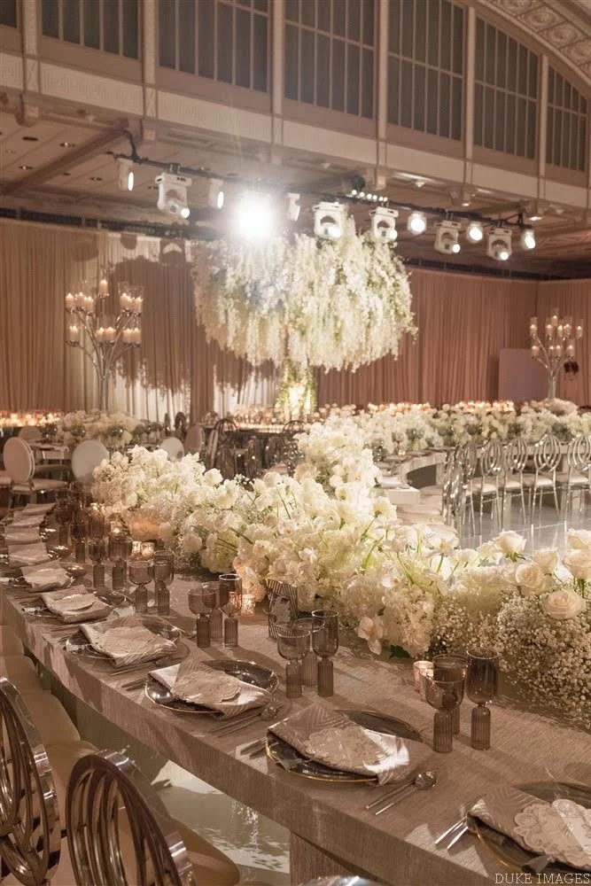 Elegantly decorated banquet hall with long table set for guests, featuring white floral centerpieces, silverware, napkins, and candles, with chandeliers and draped curtains in the background.