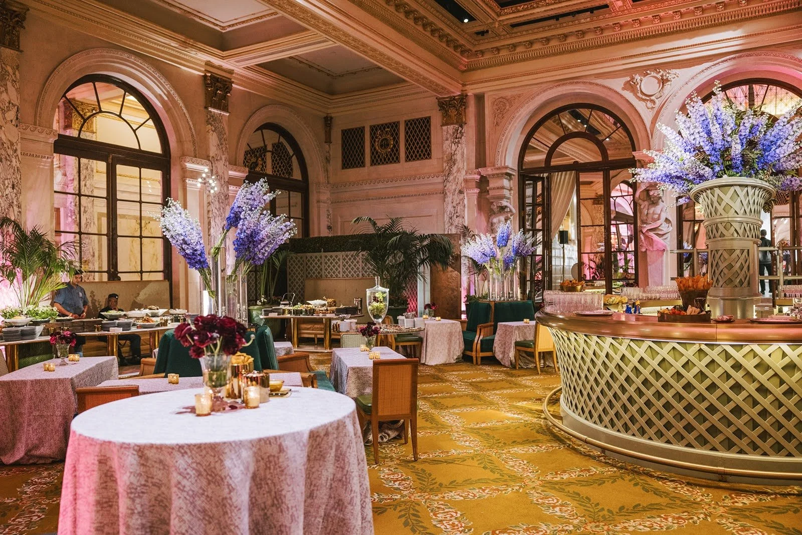 Elegant banquet hall decorated with tall purple flowers, round tables with pink tablecloths, and a curved bar area with large floral arrangements, ornate architecture, and large windows.