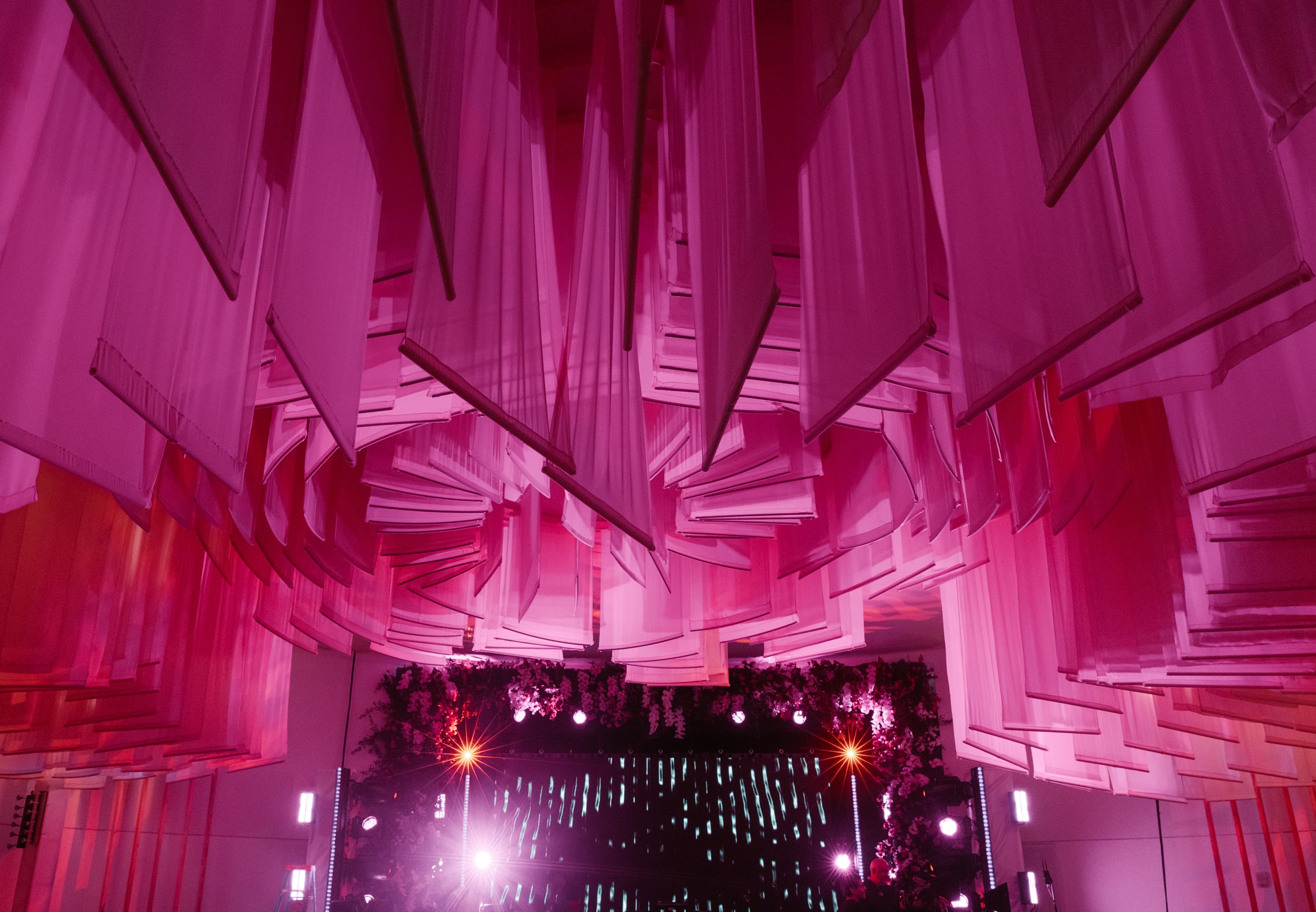 Interior view of a conference or event hall with suspended pink fabric panels and pink lighting, a stage with LED screen, and floral decorations.