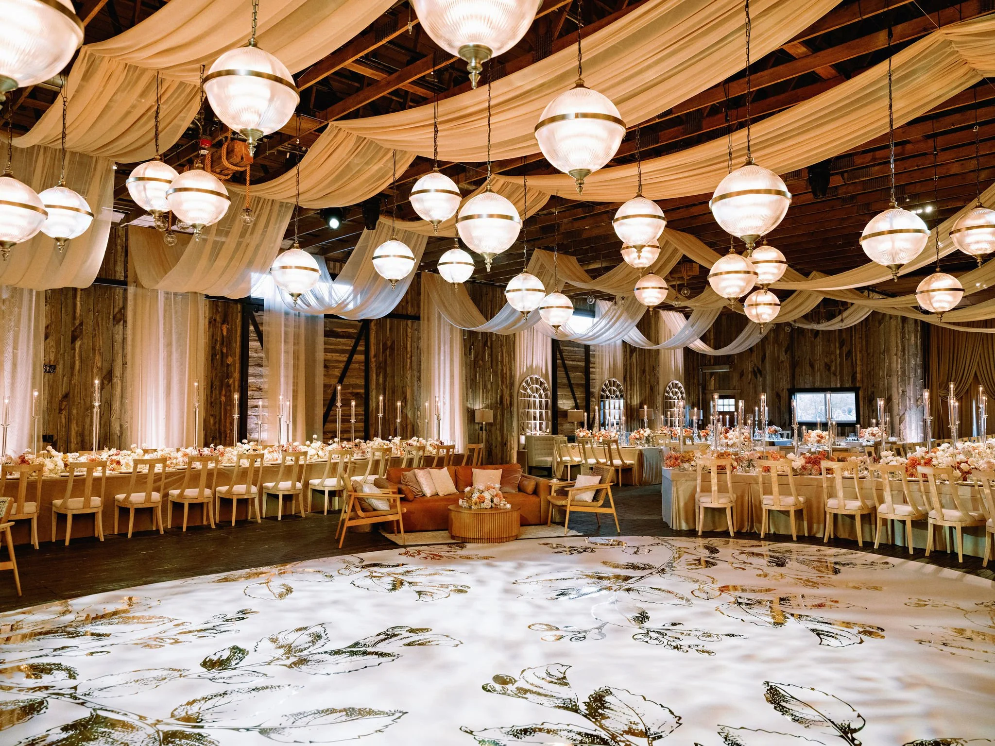 An elegant event space with wooden walls, draped fabric ceiling, and hanging spherical pendant lights. Round tables decorated with floral arrangements and chairs surround a dance floor with a leaf design.
