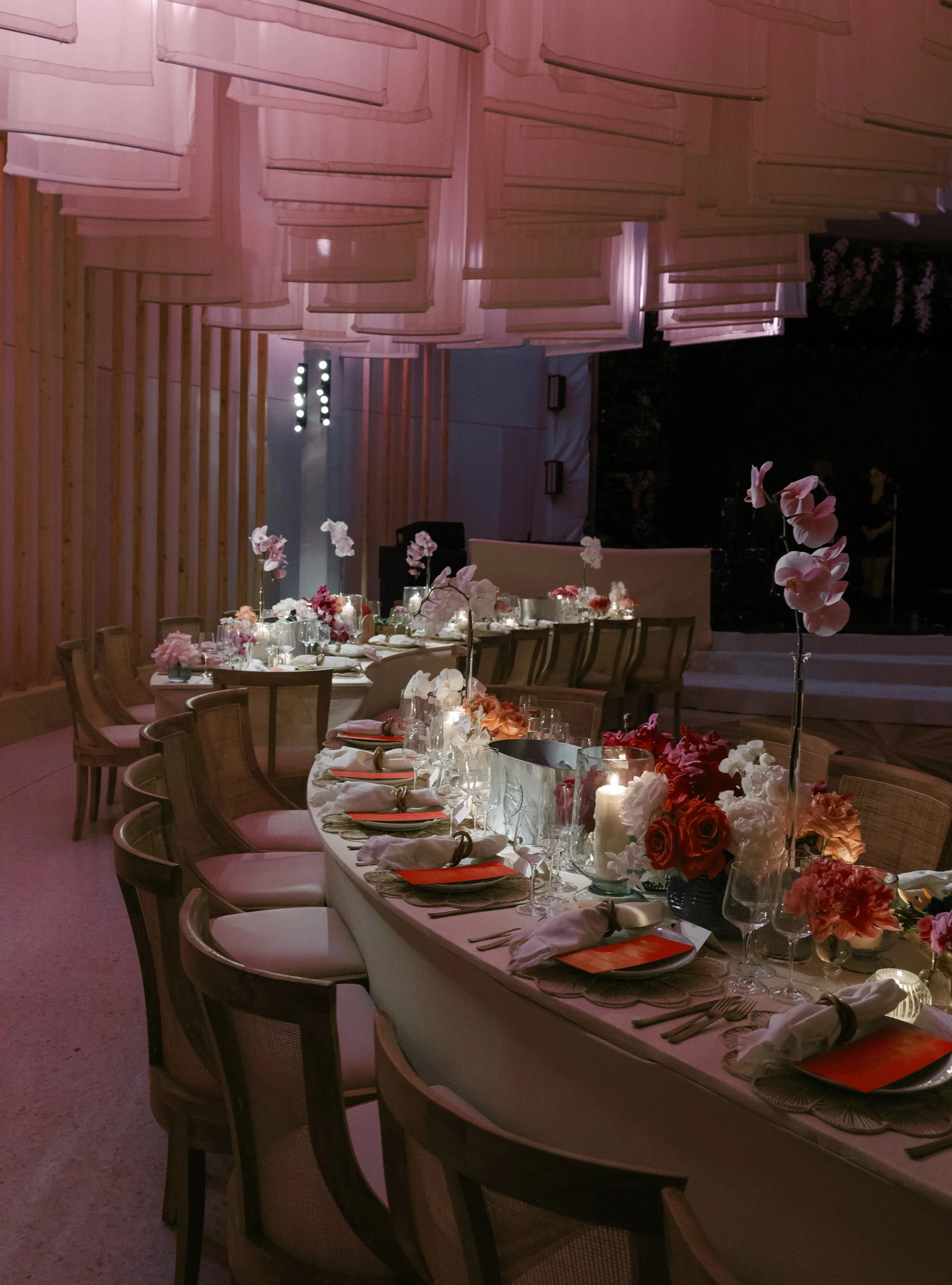 Elegant, softly lit banquet table decorated with pink and white flowers, candles, and tableware, set for an event in a modern venue with wooden wall panels and a stage in the background.