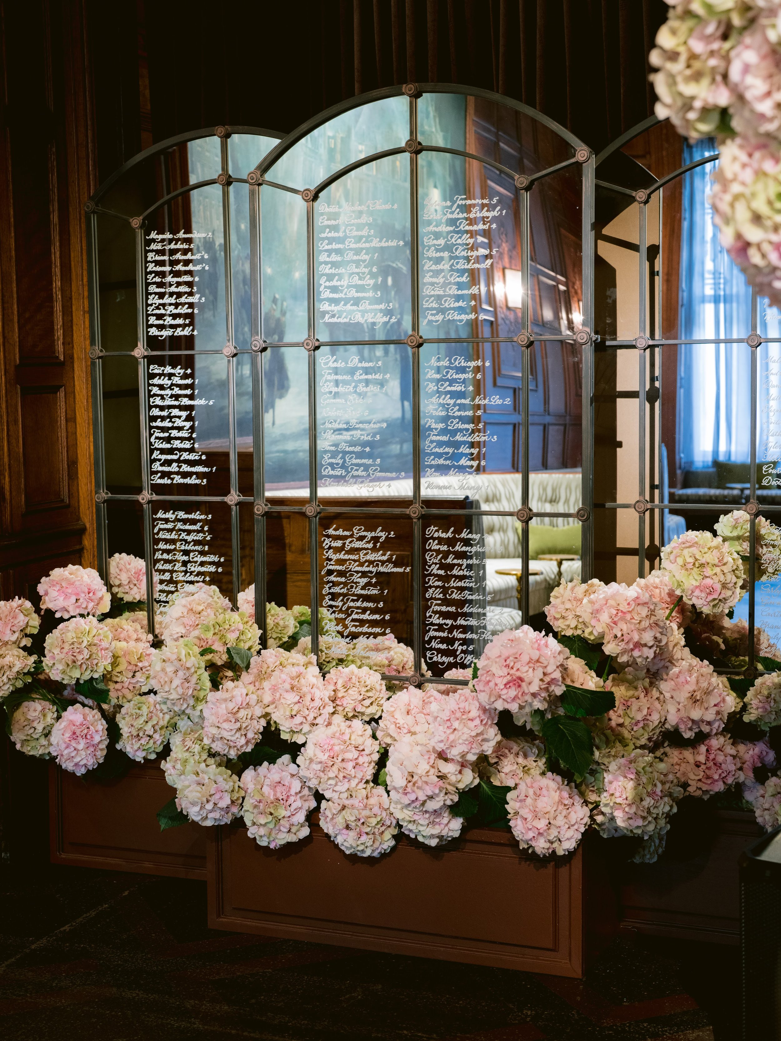 Detailed seating chart with names written in white cursive on glass panels, surrounded by pink and white hydrangea flowers, set against a wooden interior with windows and curtains.