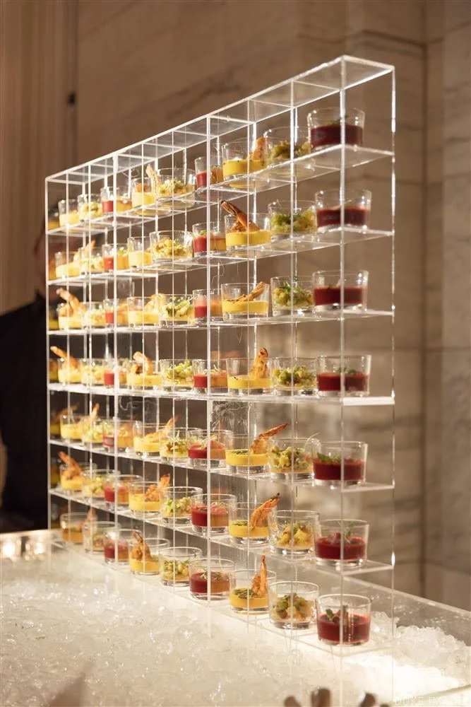 Multiple small, clear cups filled with colorful appetizers are displayed on a transparent, multi-tiered, acrylic stand at an event or party.