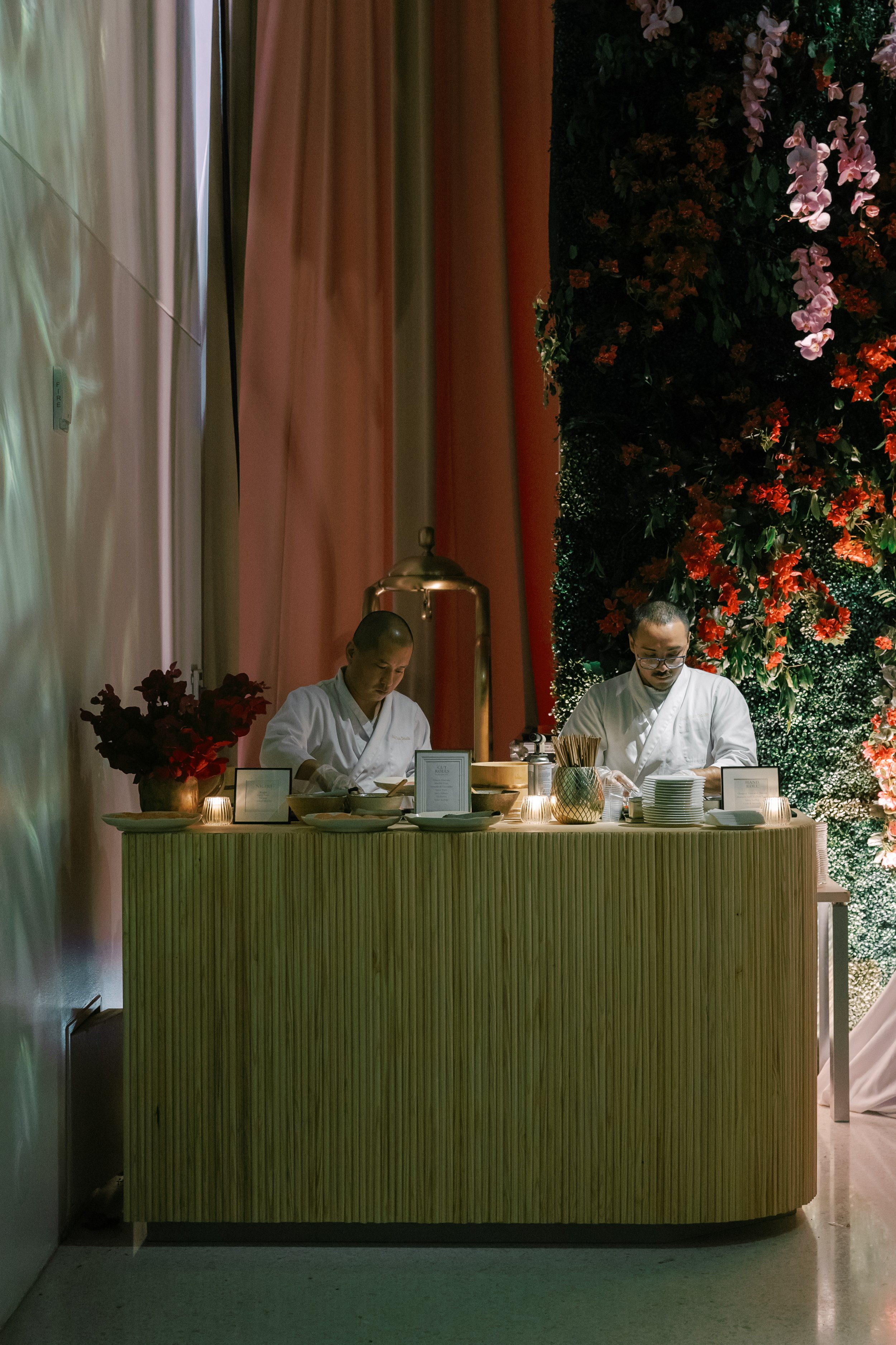 Two chefs preparing food at a decorated buffet table with floral backdrop and candles.