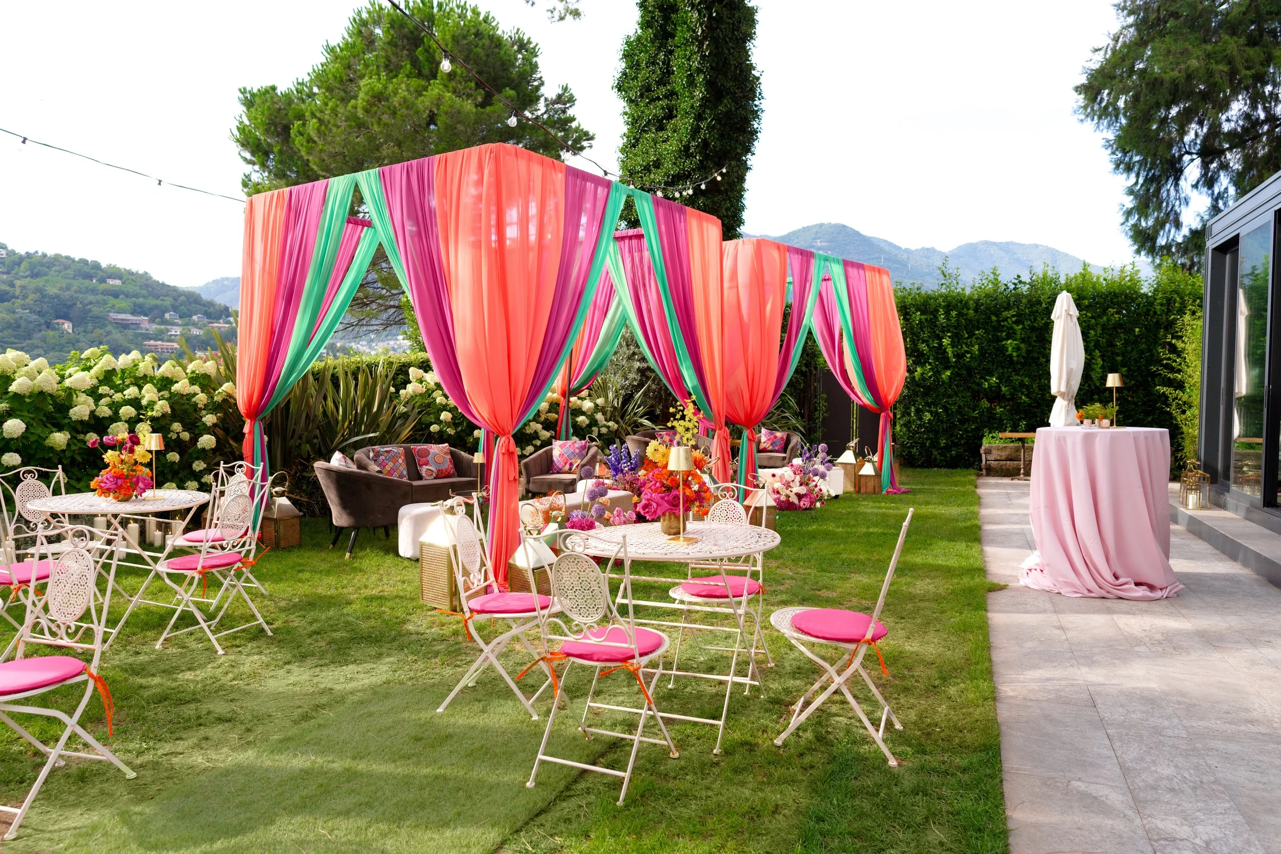 Colorful outdoor event setup with pink and teal draped fabric forming a canopy, surrounding seating with pink cushions, white tables with floral centerpieces, and a pink draped high-top table on a patio. Green trees and hills in the background.