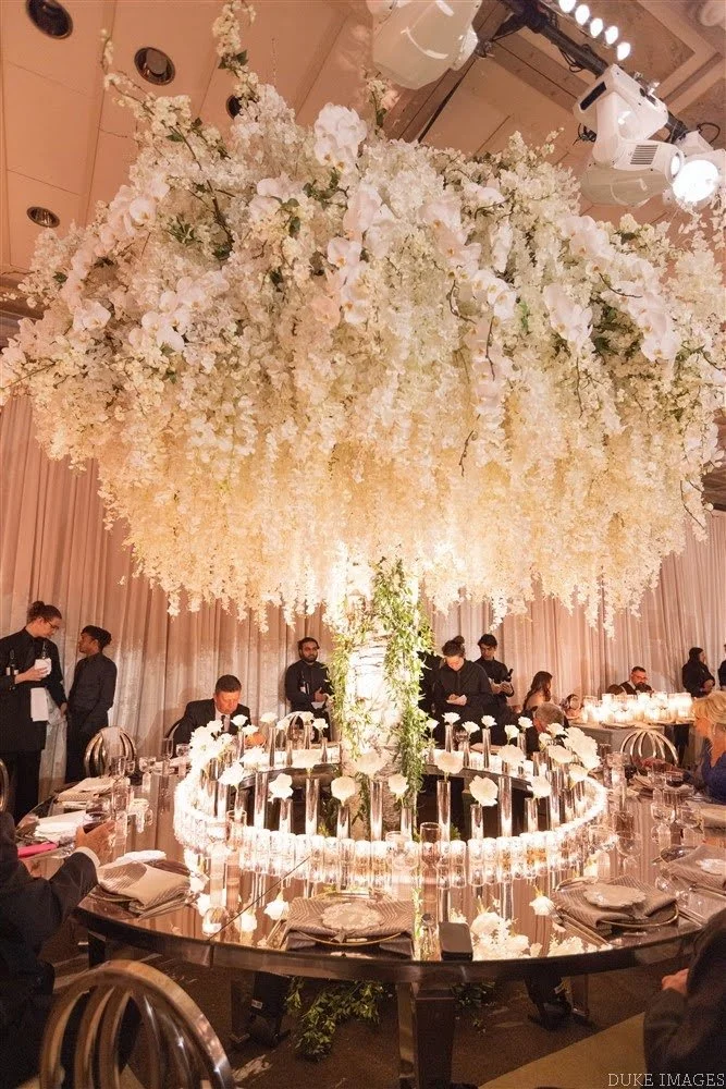 Overhead view of an elegant round banquet table with a large floral chandelier hanging above. The table is set with plates, silverware, napkins, and glassware, with a circular glass candle holder centerpiece. The floral chandelier is made of white fl