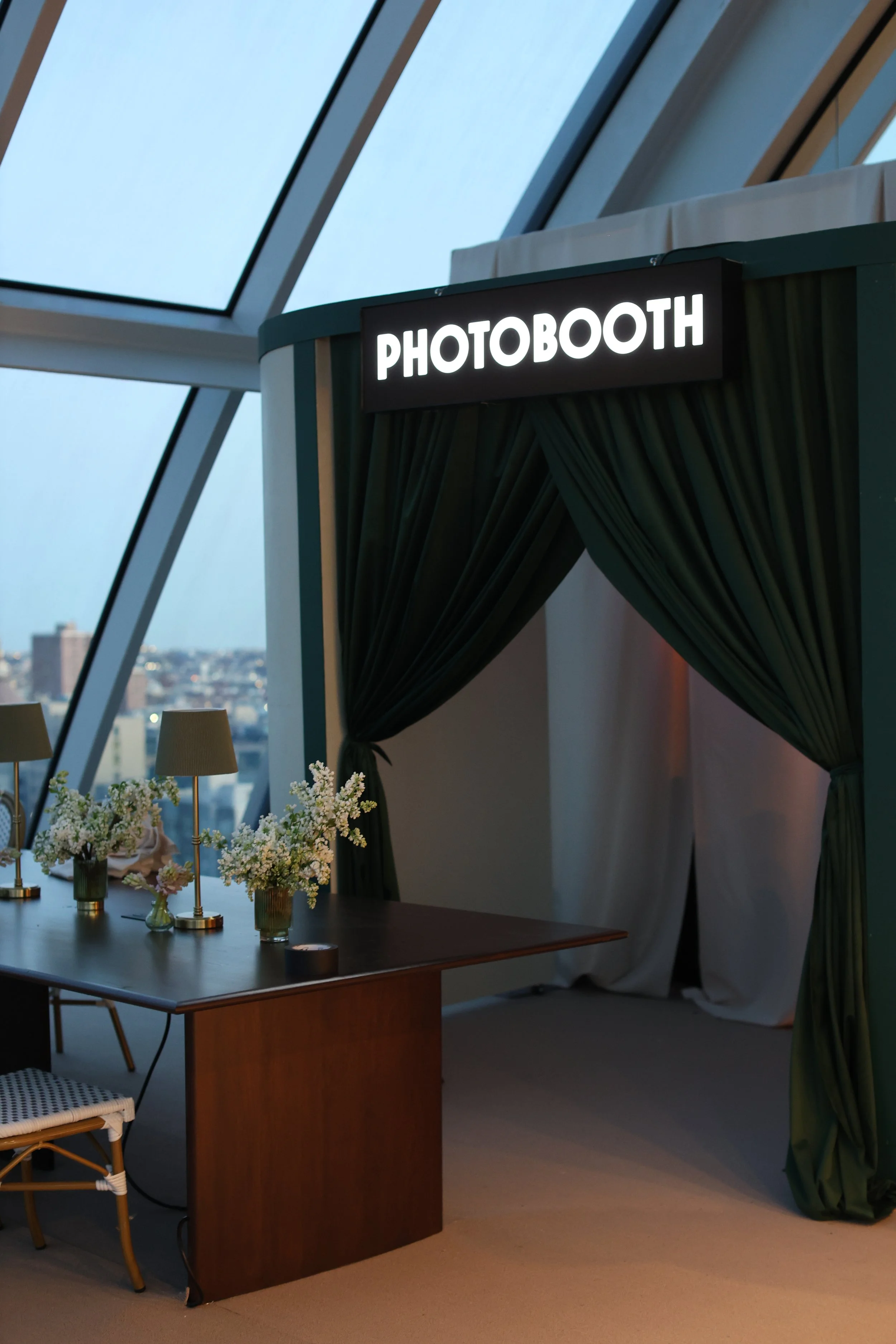 An indoor photo booth area labeled 'Photobooth' with dark green curtains, a wooden table decorated with small lamps and flowers, and a city skyline visible through large glass windows in the background.