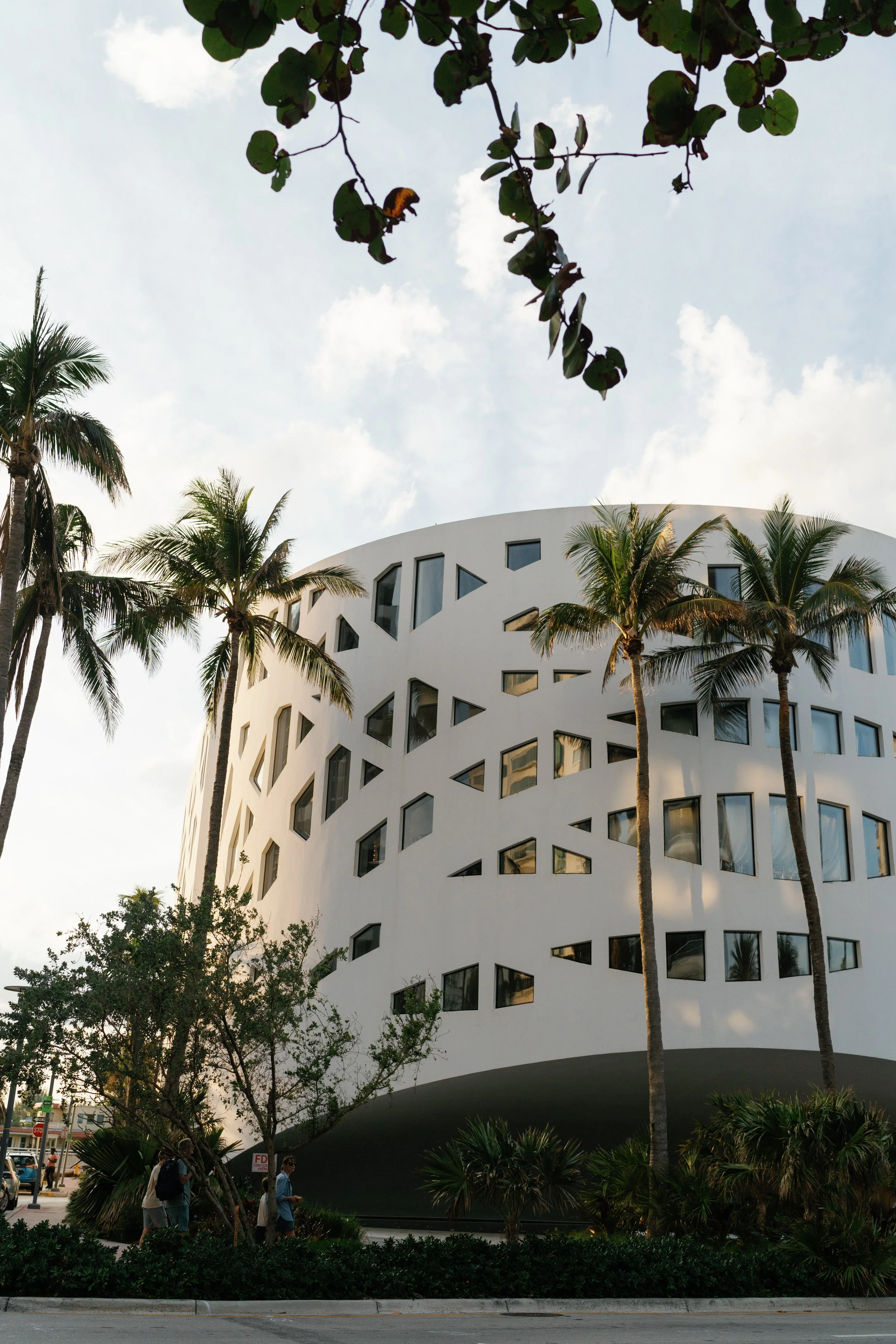 Modern white building with geometric windows, surrounded by tall palm trees and greenery, with a cloudy sky overhead.