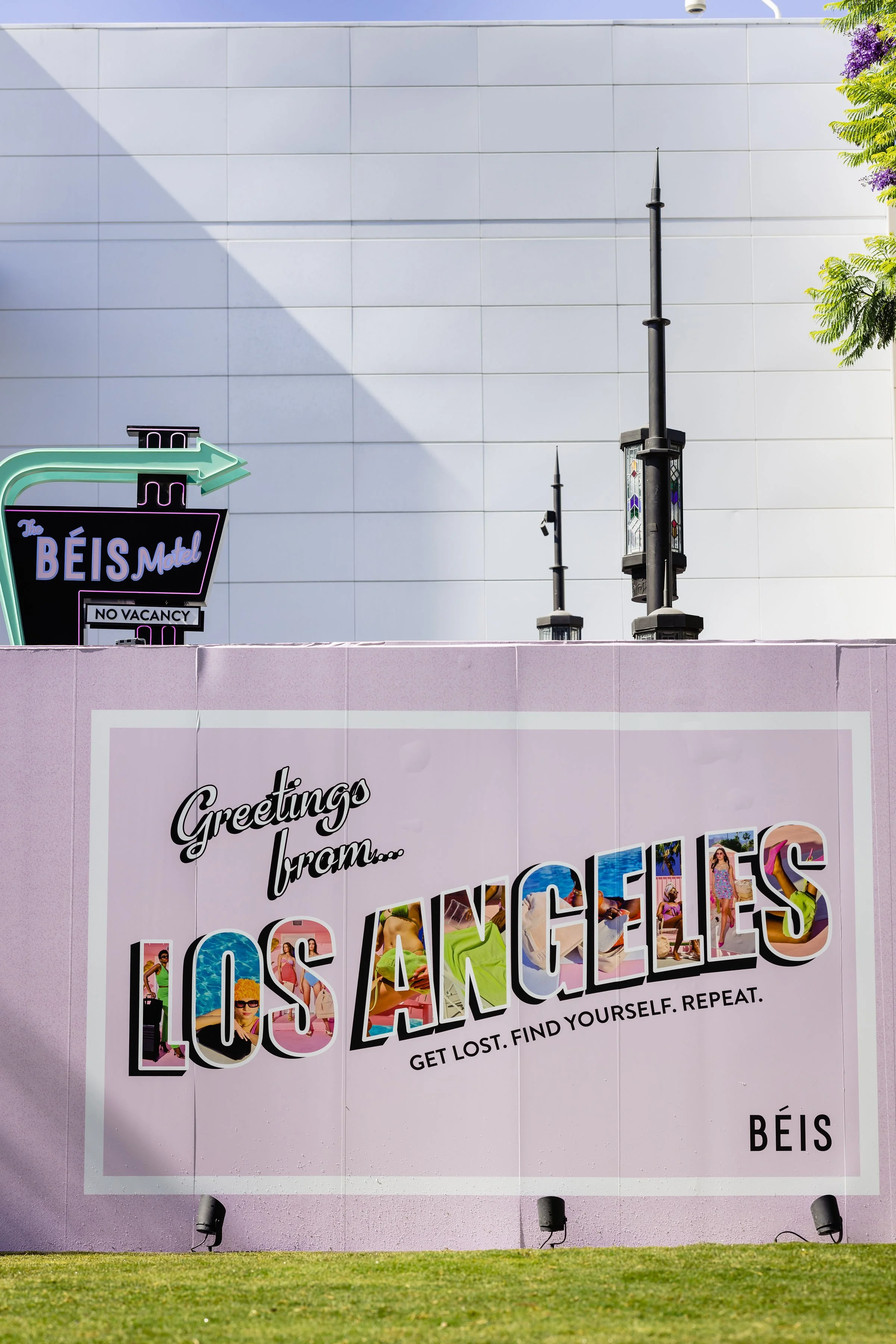 A large pink billboard with the text "Greetings from... Los Angeles" in colorful letters filled with images of people and beaches, with the tagline "Get lost. Find yourself. Repeat." and the word "Béïs" at the bottom. Part of a purple neon sign for t