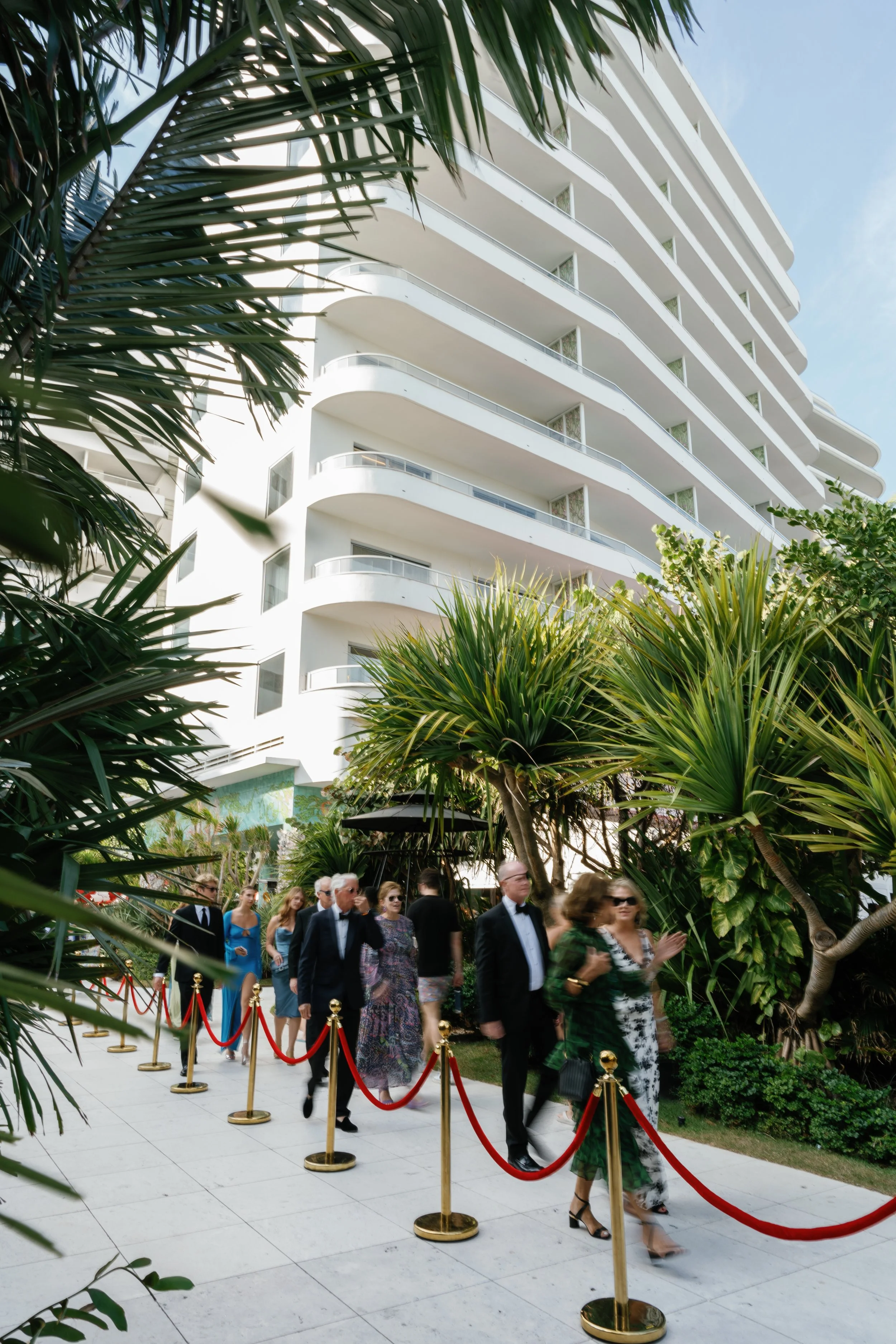People dressed in formal attire walking along a sidewalk lined by red velvet ropes, with a modern white building and lush greenery in the background.
