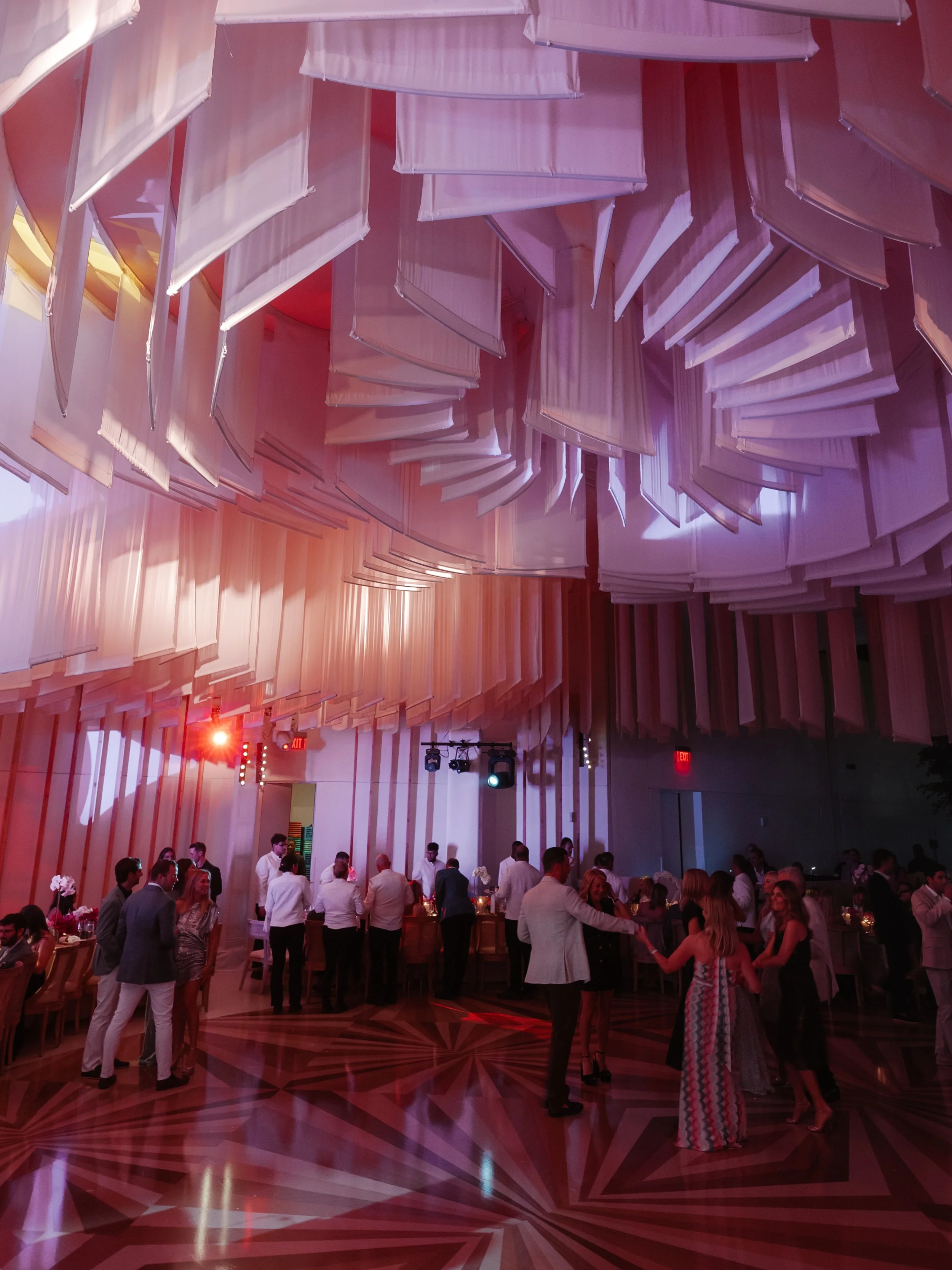 People dancing and socializing at a formal event or wedding reception in a decorated ballroom with geometric patterned floor and hanging fabric decor from the ceiling.