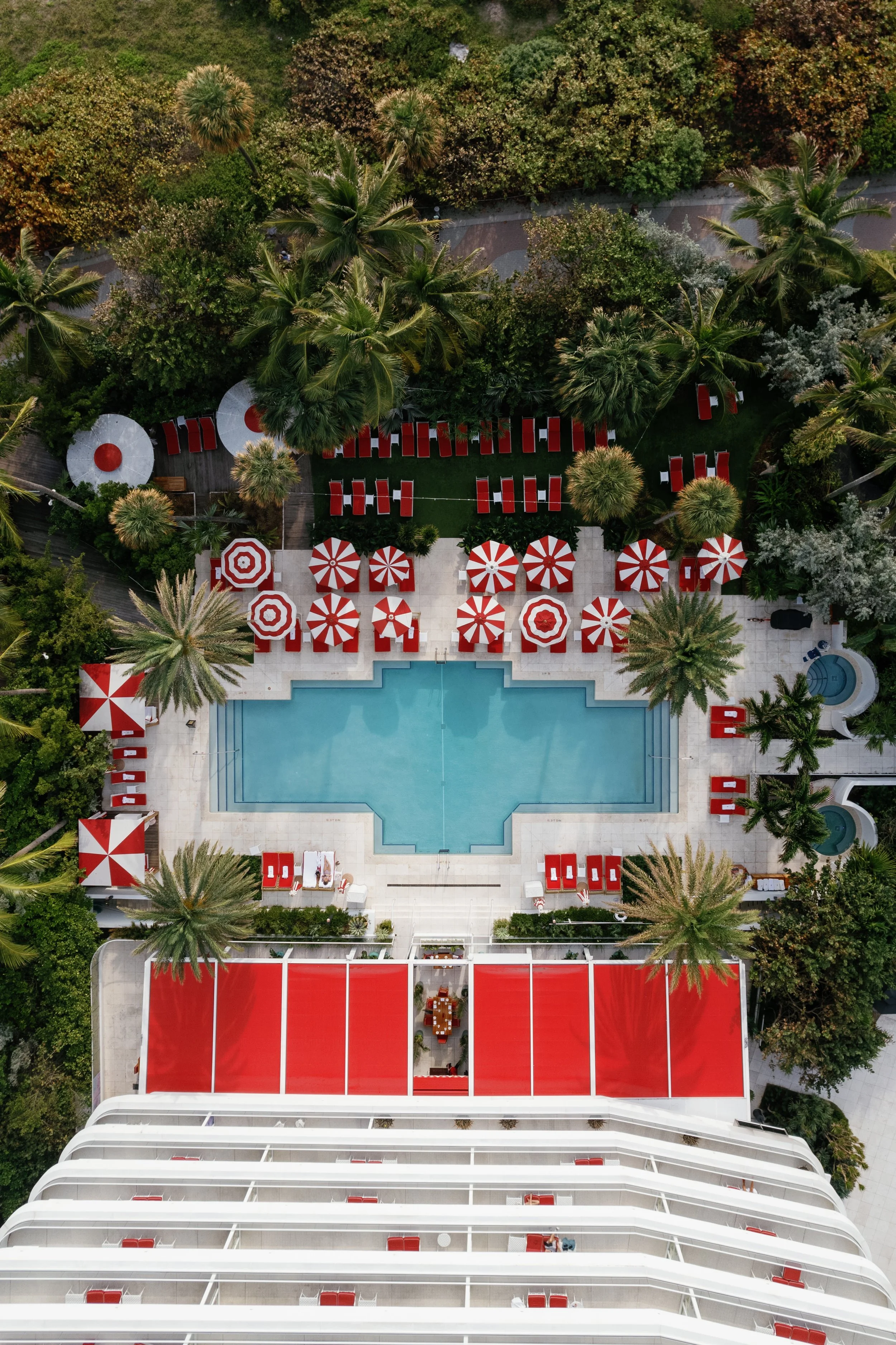 An aerial view of a hotel pool area with red and white striped umbrellas, palm trees, lounge chairs, and lush greenery surrounding the space.