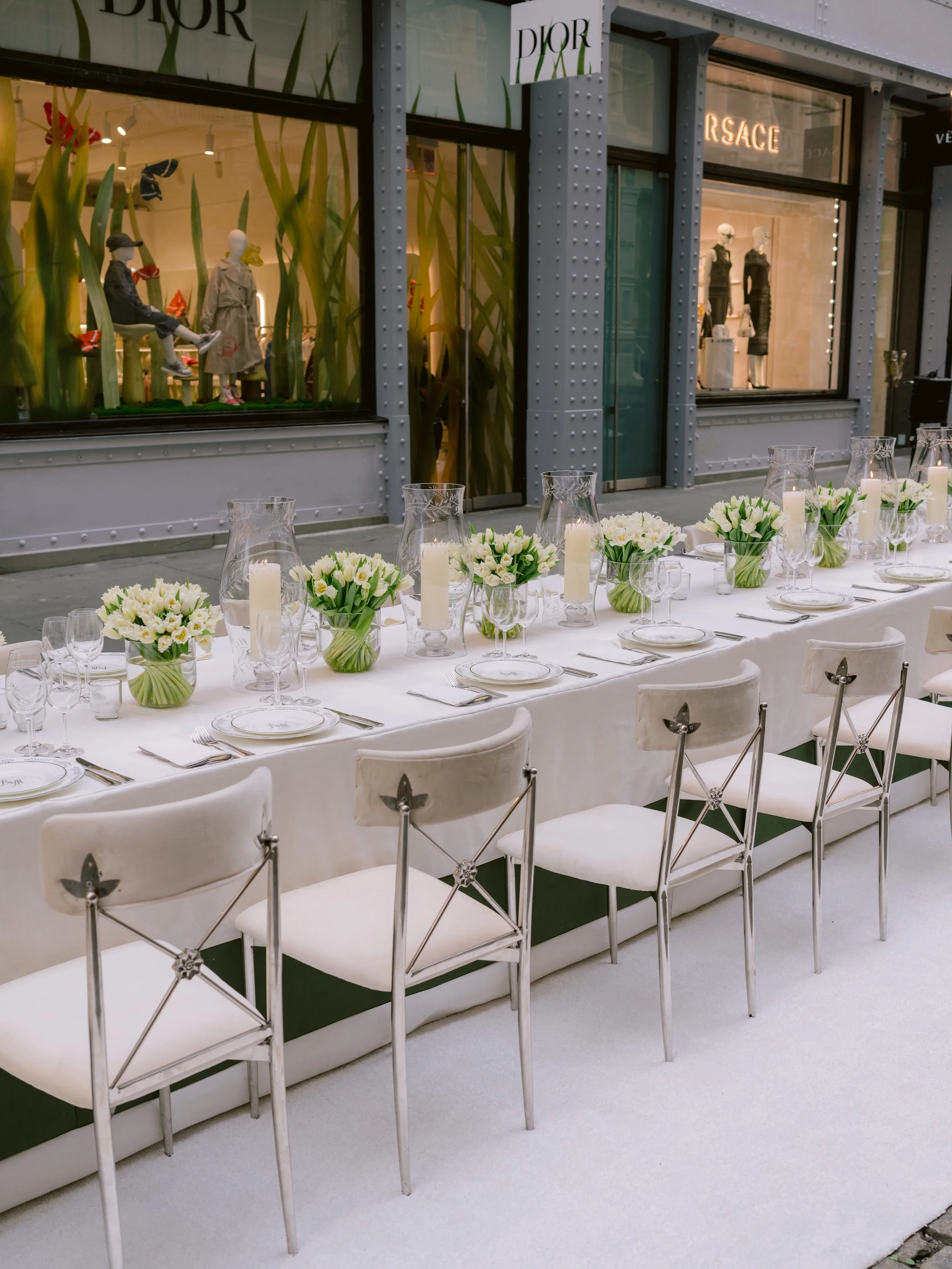 Elegant outdoor dining setup with a long table covered with a white tablecloth, decorated with white flower centerpieces, glass candles, and fine glassware, set in front of a storefront window displaying mannequins dressed in designer clothing and la