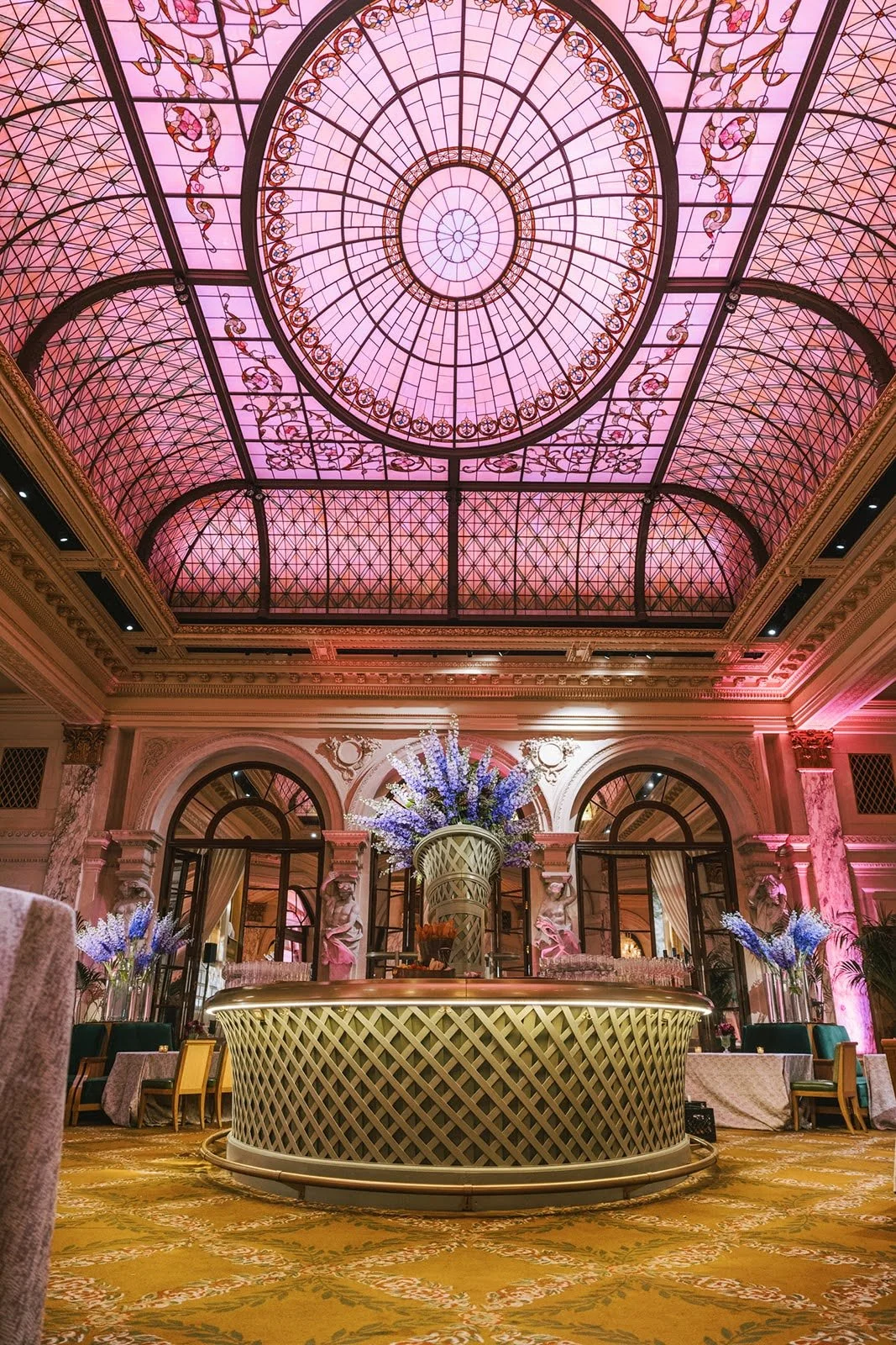 A luxurious hotel lobby featuring a large ornate pink stained glass ceiling with intricate designs. The space has elegant arches, marble columns, and crystal chandeliers. The center of the room has a round counter with a tall floral arrangement of pu