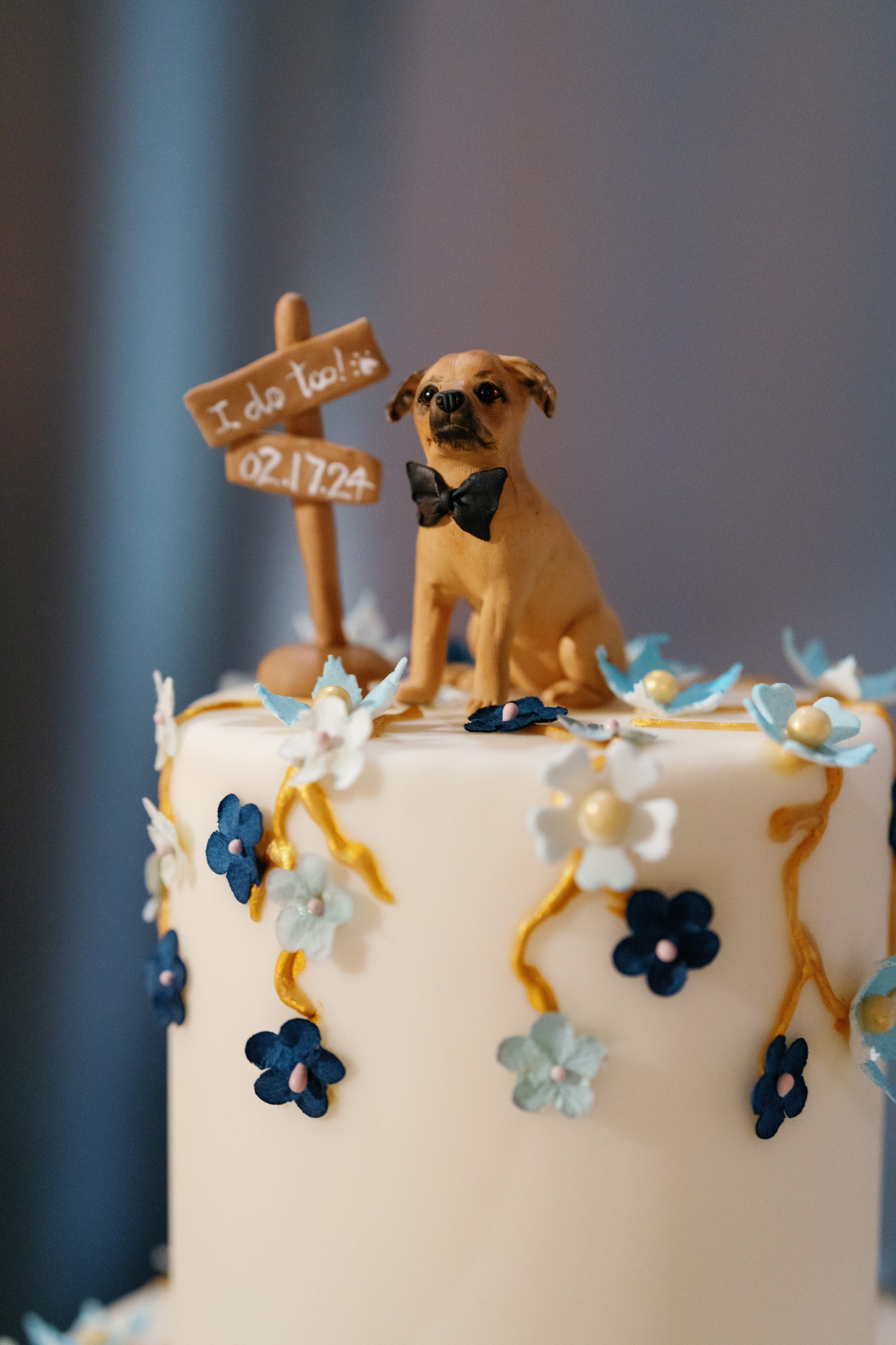 A wedding cake decorated with white and blue flowers and a small dog figurine wearing a black bow tie on top. The figurine is sitting in front of a small sign that reads 'I do too!' with a paw print, along with a date '02/17/24.'